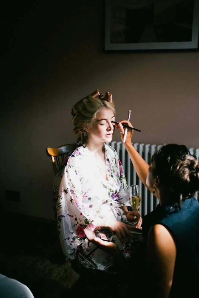 A woman sitting with her eyes closed, wearing a floral robe, has her makeup done by a makeup artist holding a makeup brush and a palette. She is holding a glass of champagne. The scene is indoors with a dark wall and framed picture above.
