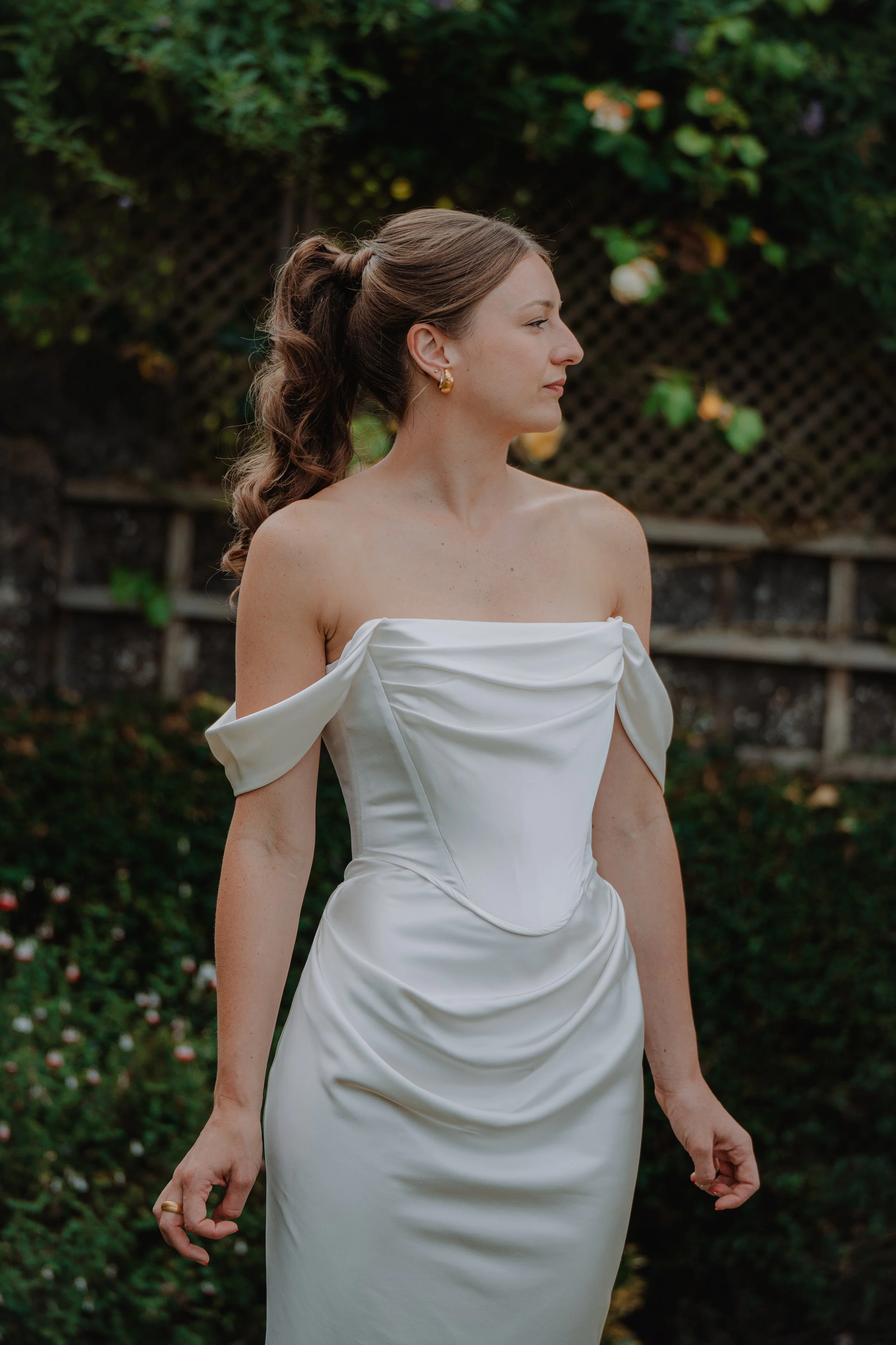 A woman wearing a white, off-shoulder satin wedding dress with gather details, standing outdoors in a garden with green foliage and flowers.