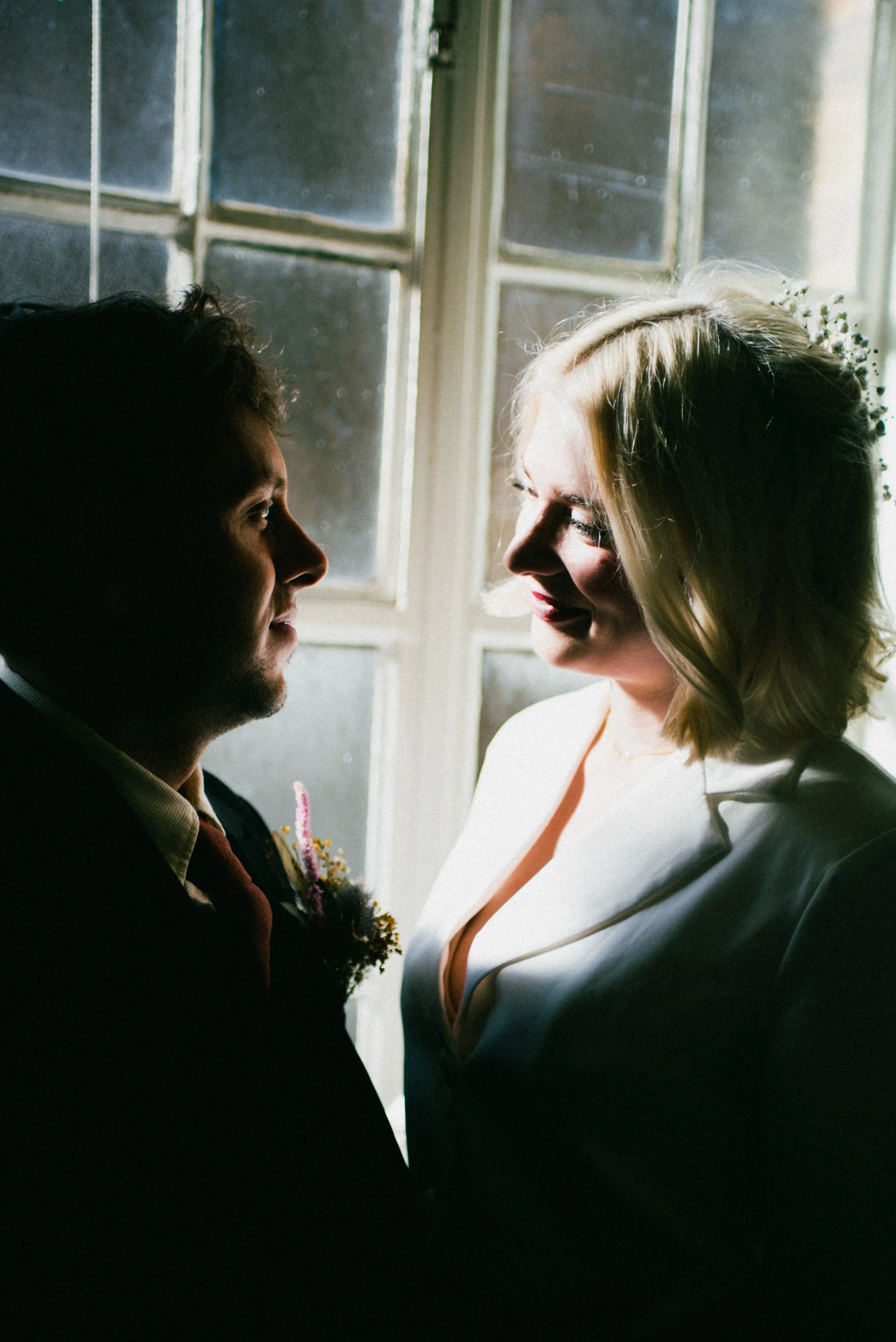 A couple looks at each other in a dimly lit room with sunlight coming through a window, highlighting their faces.