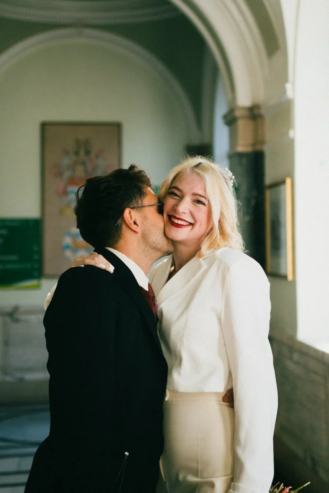 A man in a black suit kisses a smiling woman with blonde hair, wearing a cream-colored outfit, in an indoor setting with arched ceilings and framed artwork.
