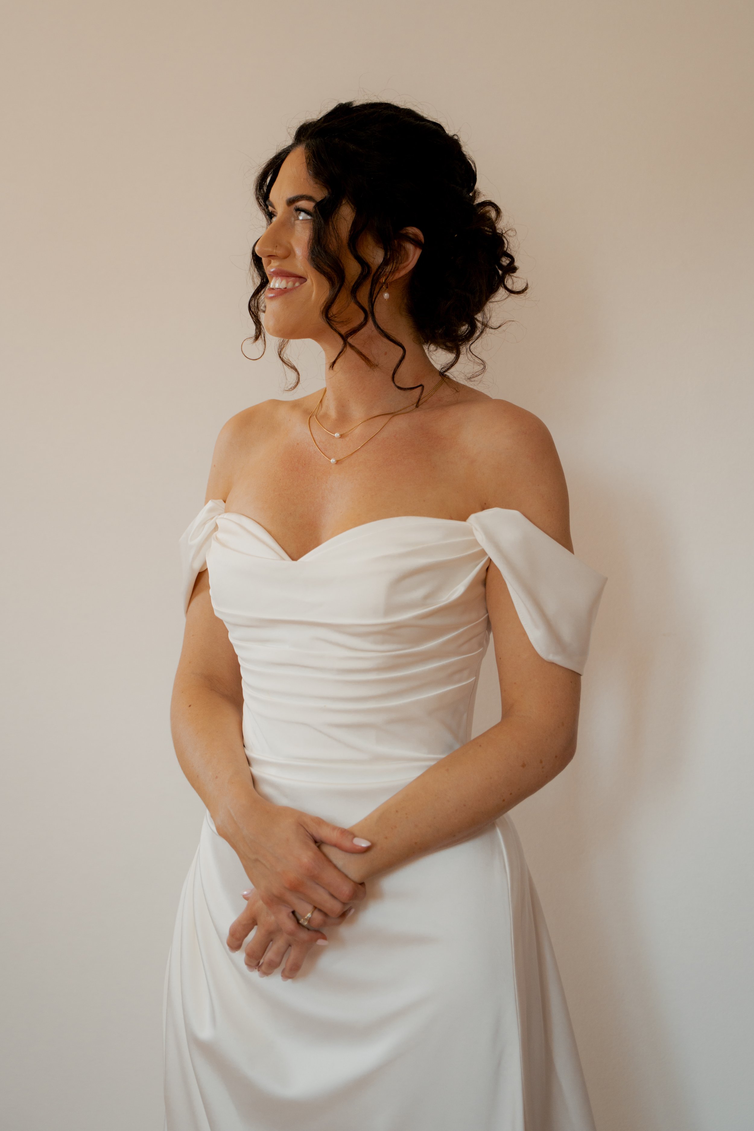 A woman in a white wedding dress with off-the-shoulder sleeves, standing sideways, smiling, with dark curly hair and wearing jewelry, against a plain beige wall.