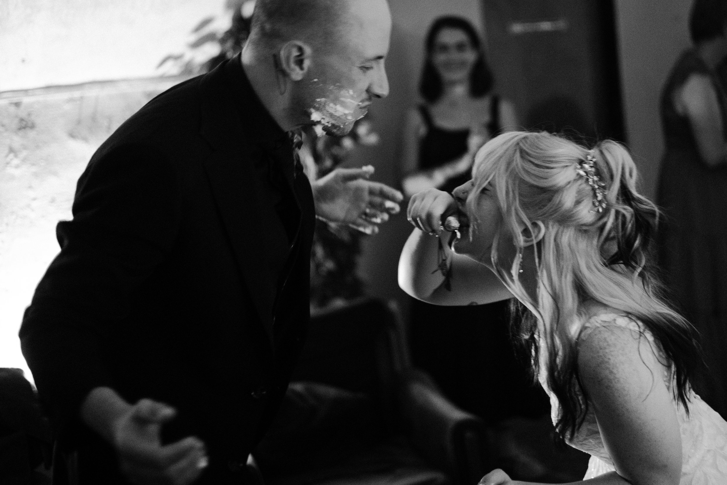A man with facial cake or frosting on his face is leaning close to a woman with long, curly hair and a decorative hairpiece, who appears to be laughing or smiling. They are at a celebration or party, with other blurred figures in the background.