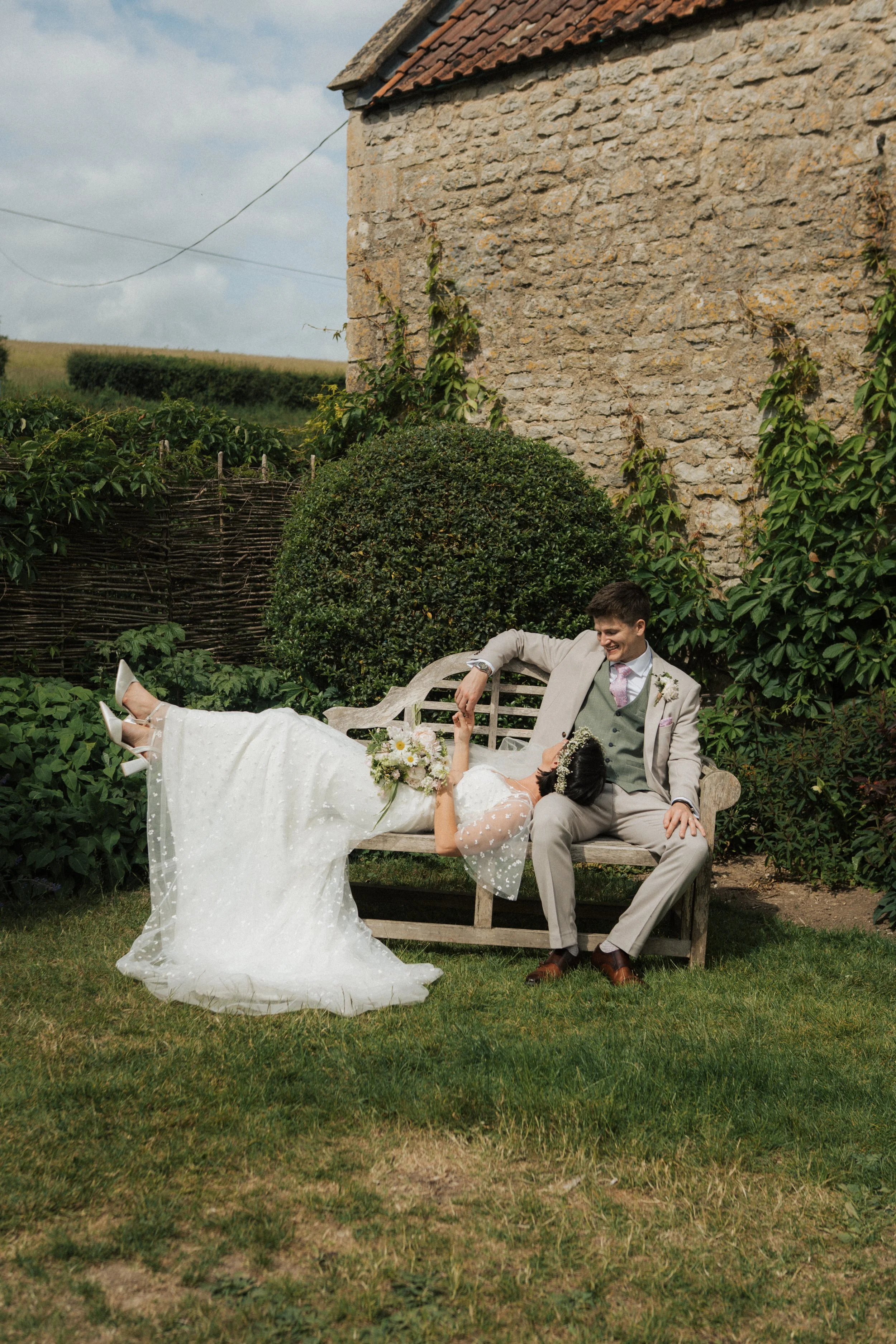 A bride and groom sitting on a bench outdoors, holding hands and smiling. The bride wears a white wedding dress and the groom wears a light grey suit. There are green bushes and a rustic stone wall in the background.