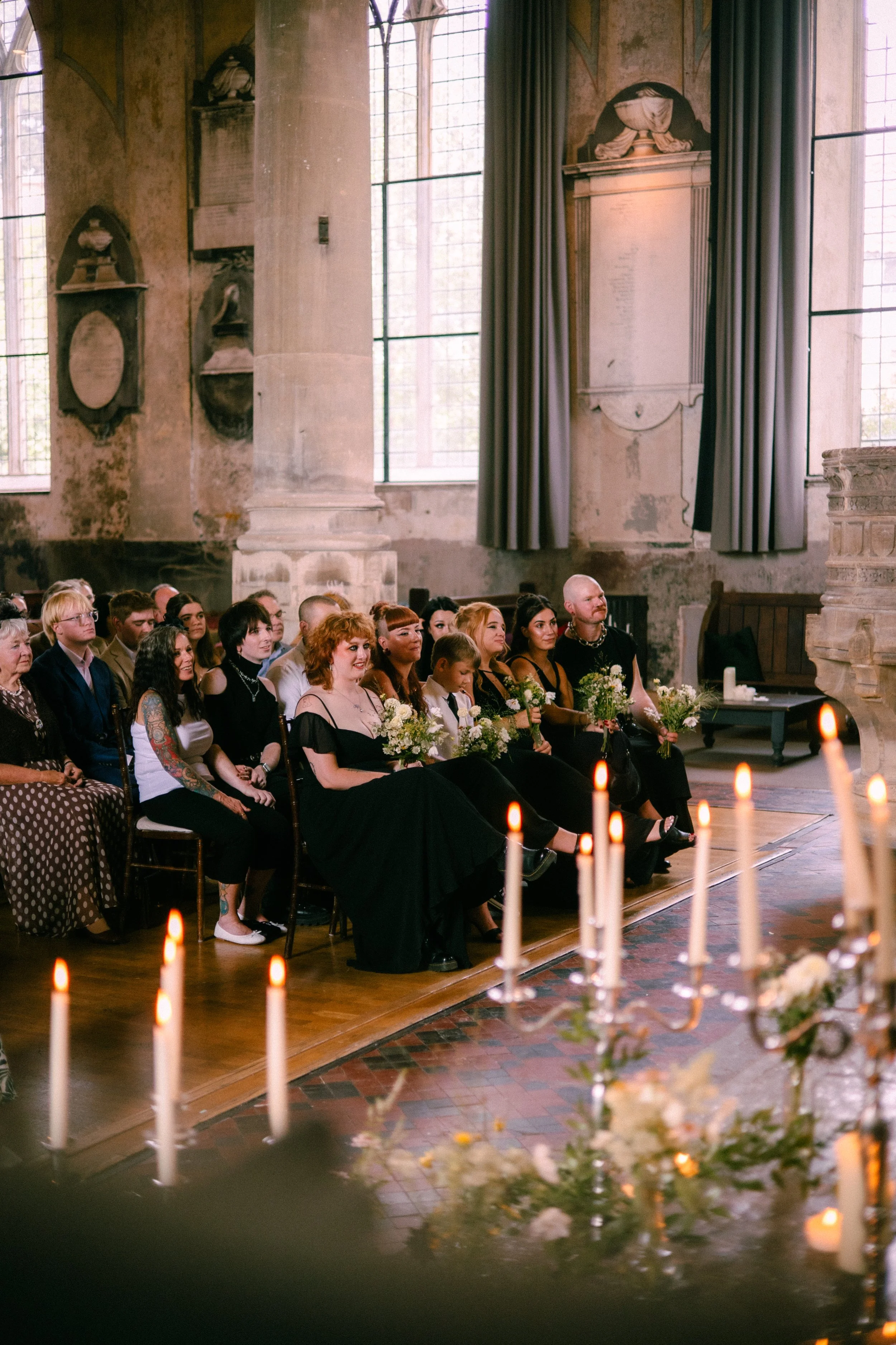 A group of people sitting in pews inside a church during an event, with candles and floral arrangements in the foreground, large windows with curtains, and decorative elements on the walls.