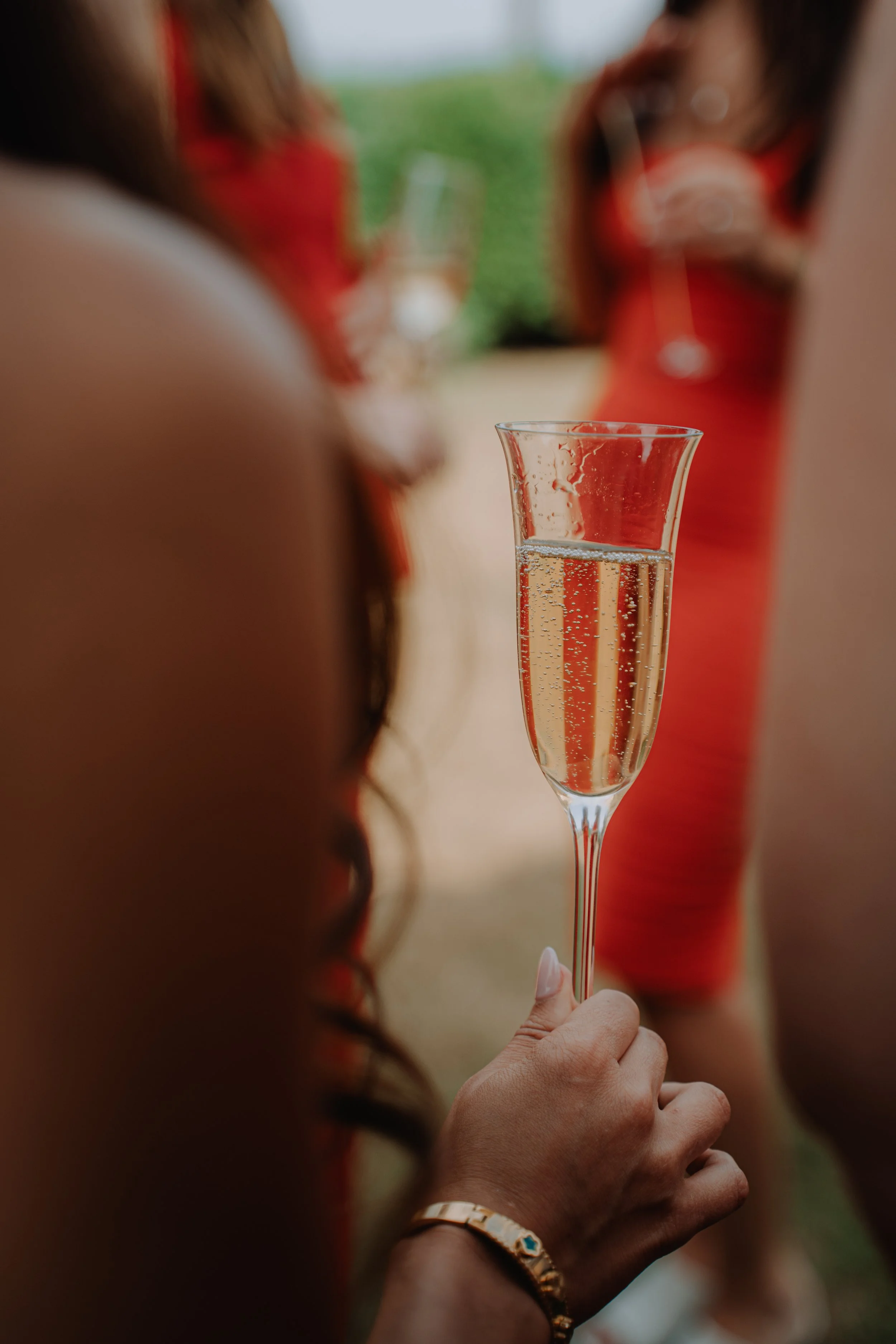 Close-up of a woman's hand holding a champagne flute filled with bubbly, with women in red dresses blurred in the background at a social gathering.