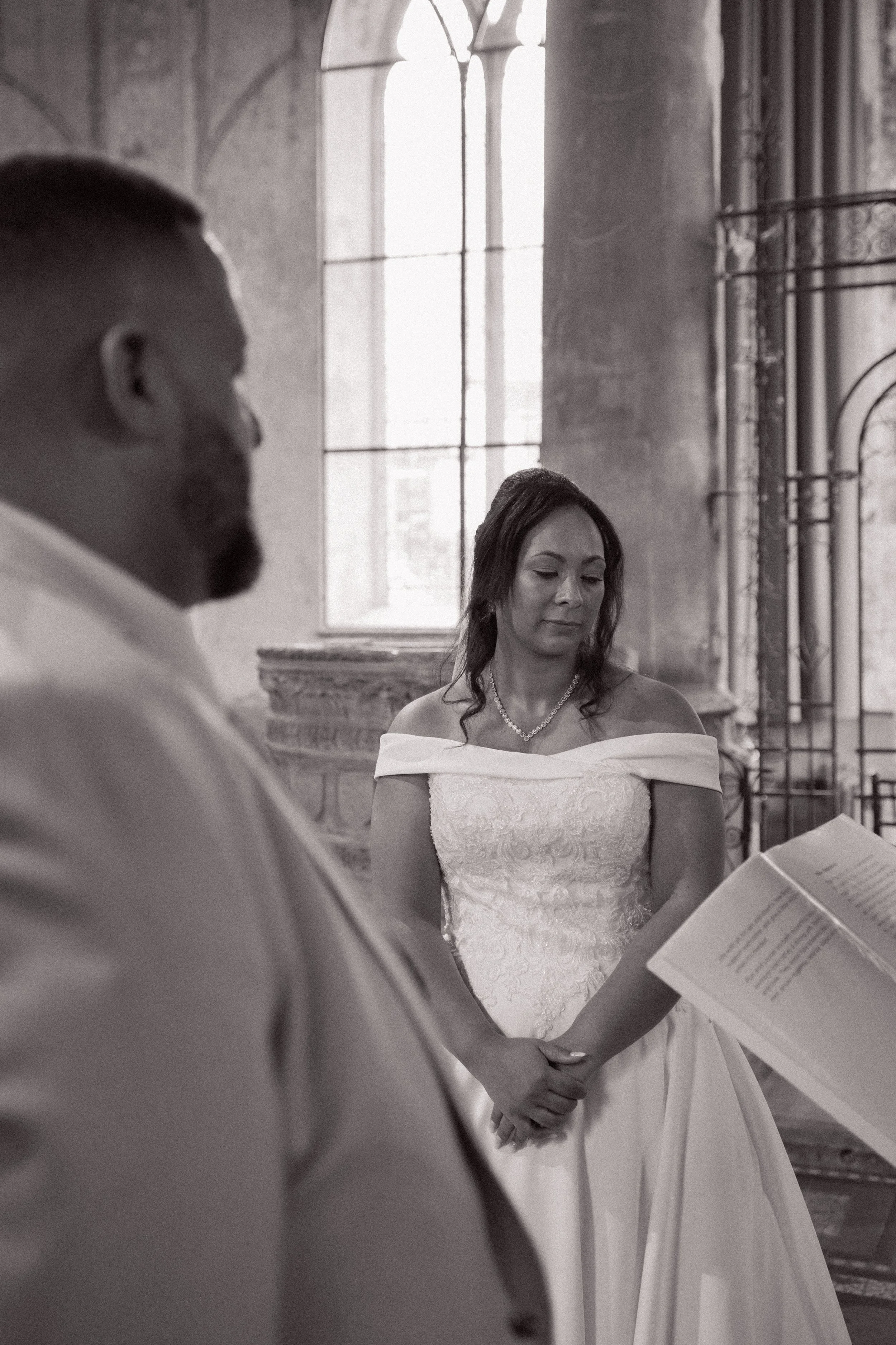 A bride and groom during a wedding ceremony in a church, with the bride in a white off-shoulder wedding dress holding her hands together and the groom slightly blurred in the foreground, as they listen to the officiant.