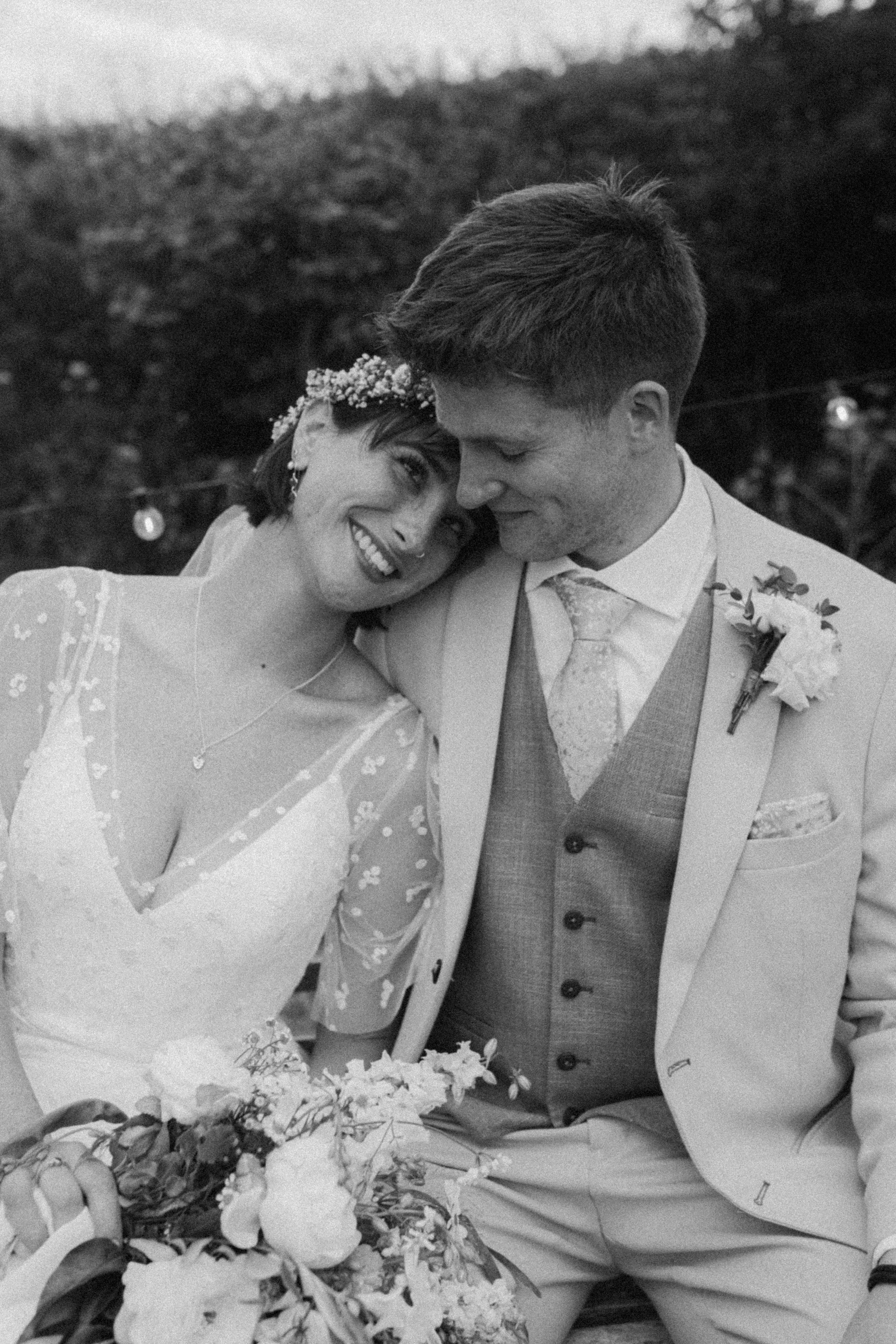 Black and white photo of a bride and groom sitting close together at their wedding, smiling, with the bride holding a bouquet of flowers.