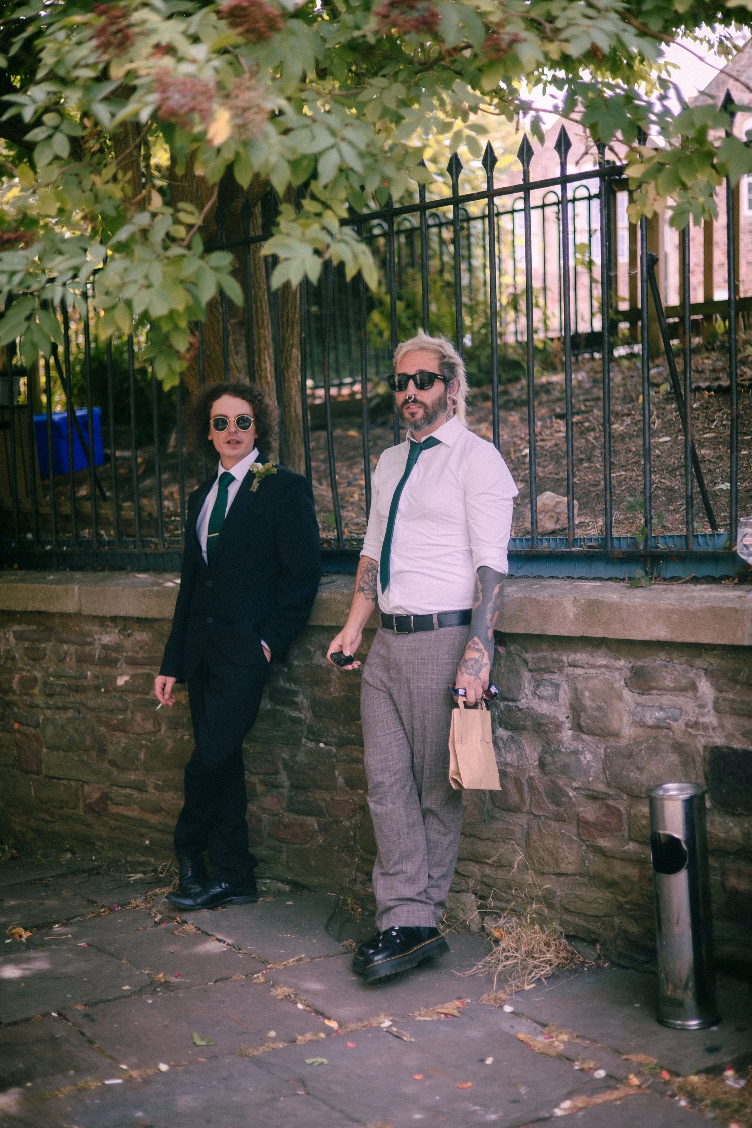 Two men standing on a sidewalk near a brick wall, both wearing sunglasses and formal attire, with trees and a black metal fence in the background.