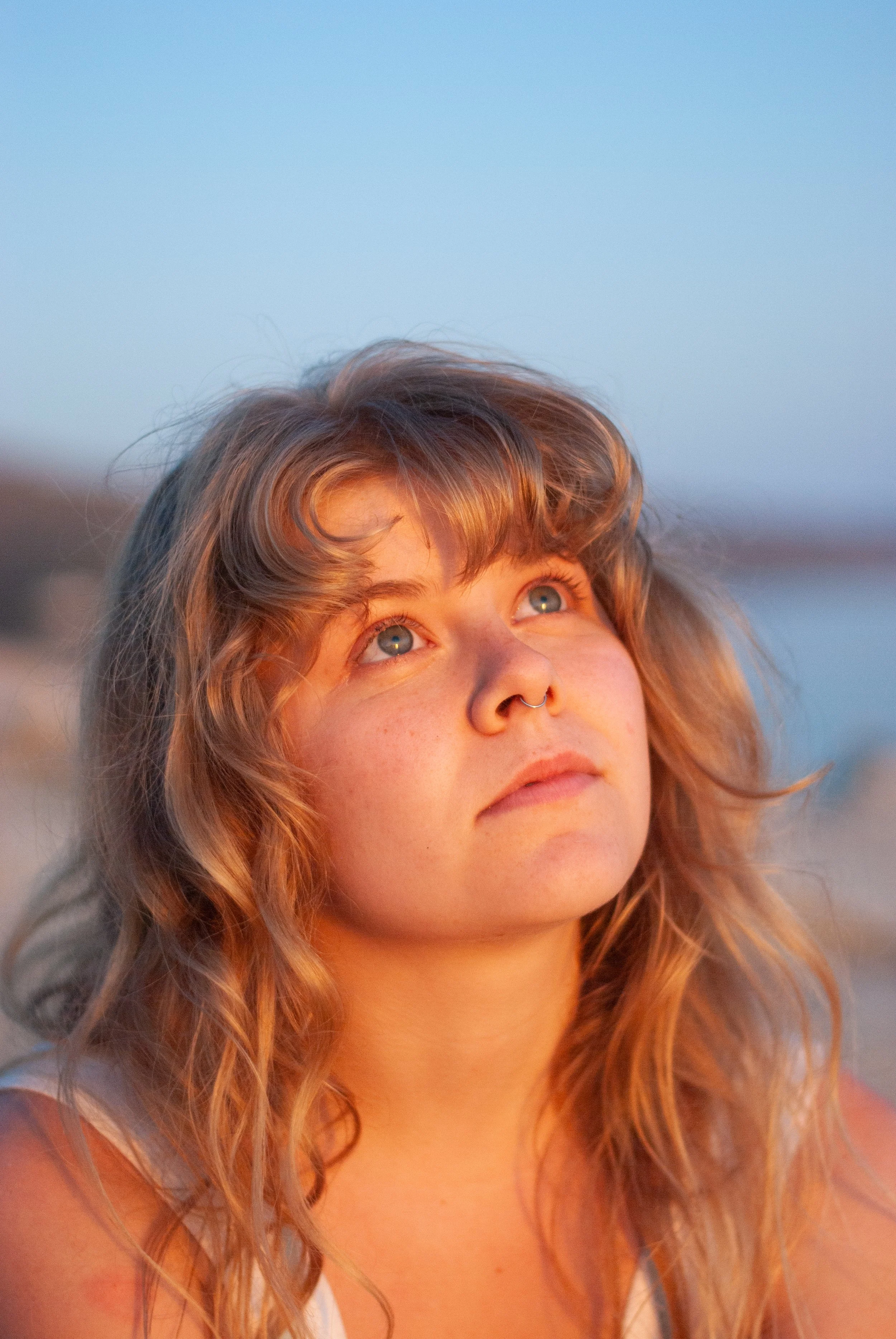 A young woman with wavy hair and a septum piercing looking up at the sky during sunset.