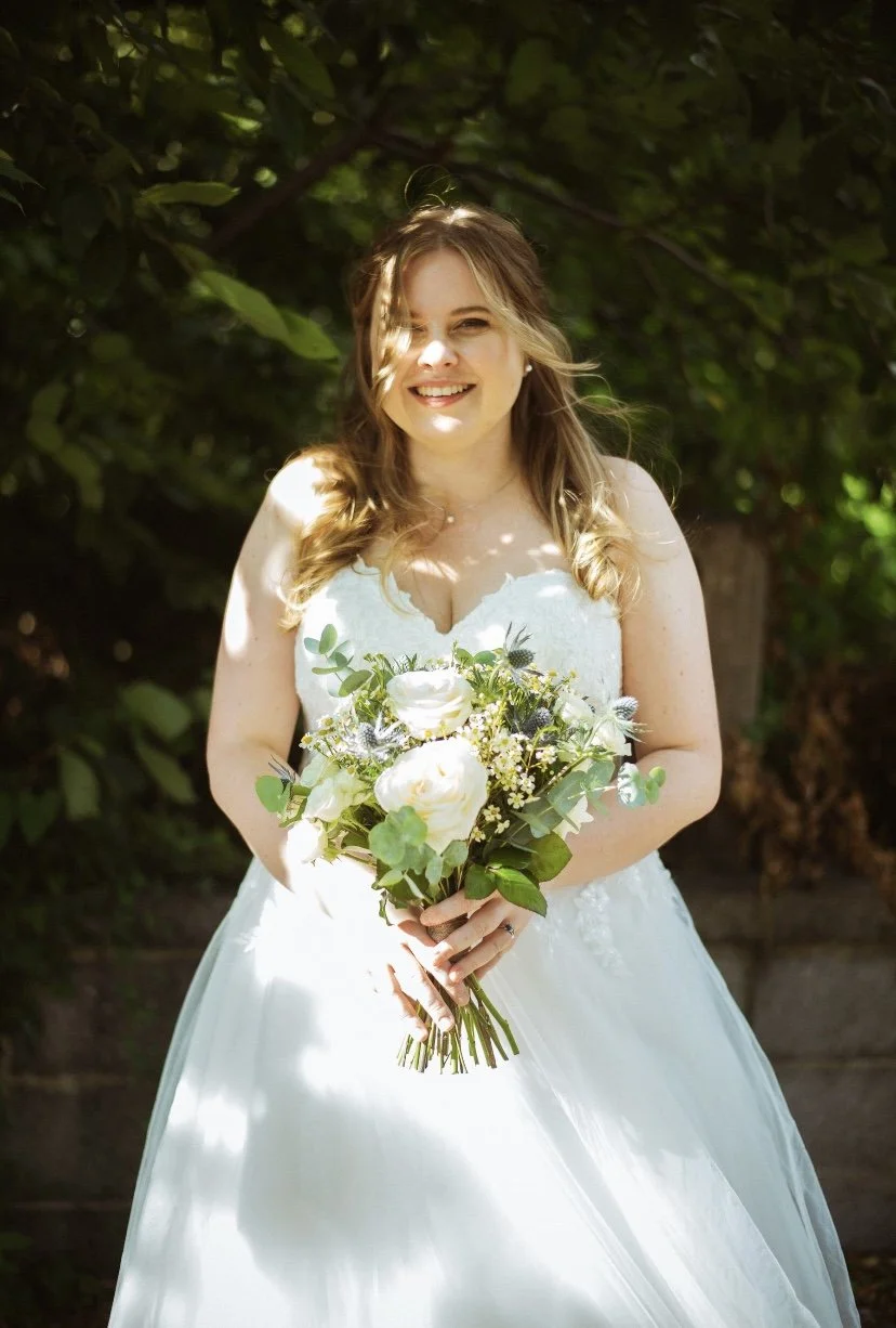 A smiling bride in a white wedding dress holding a bouquet of white and green flowers, standing outdoors with leafy green background.