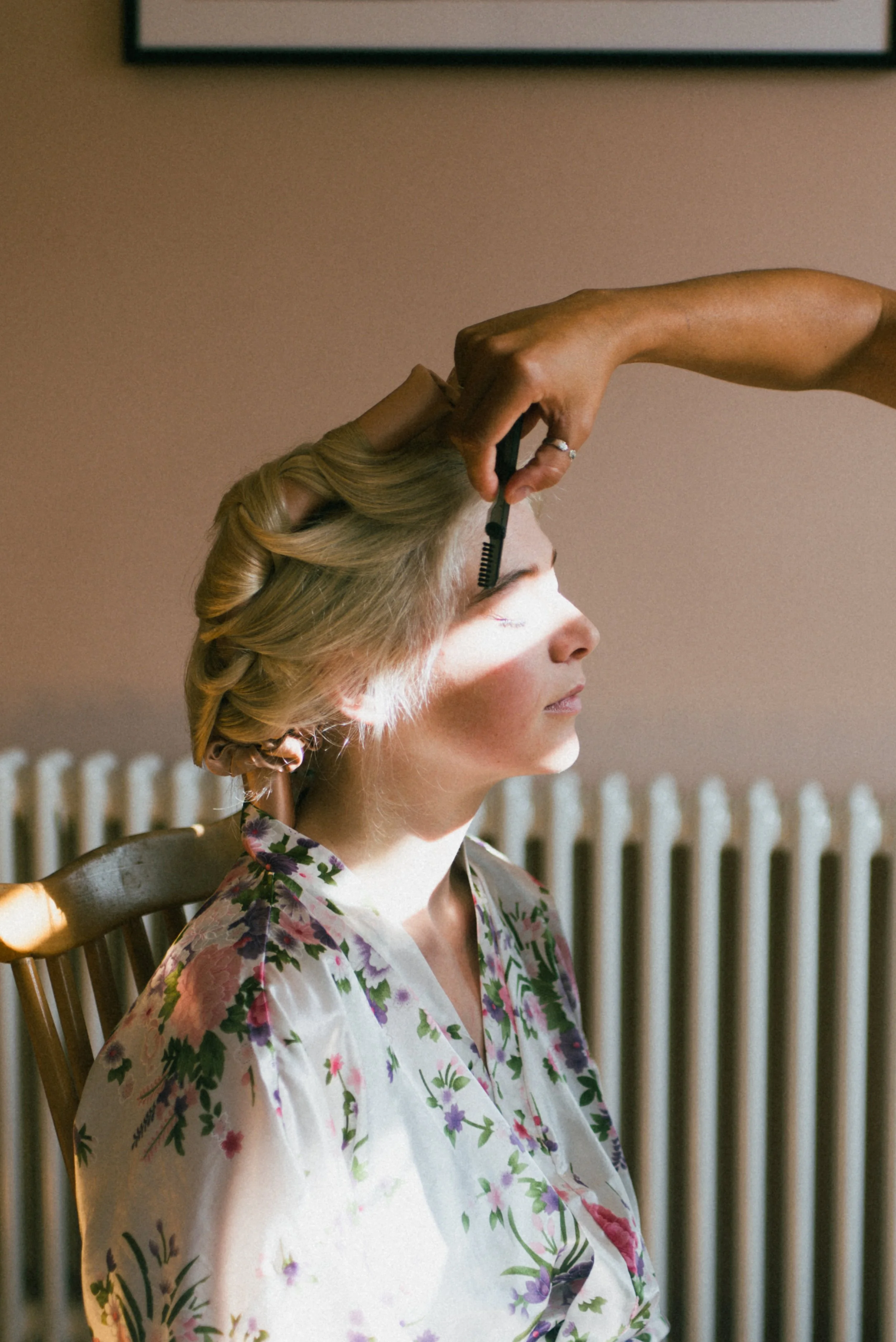 A woman with blonde hair styled in curls wears a floral robe and is getting her makeup done. A makeup artist is applying eyeshadow with a brush, with sunlight casting shadows on her face.
