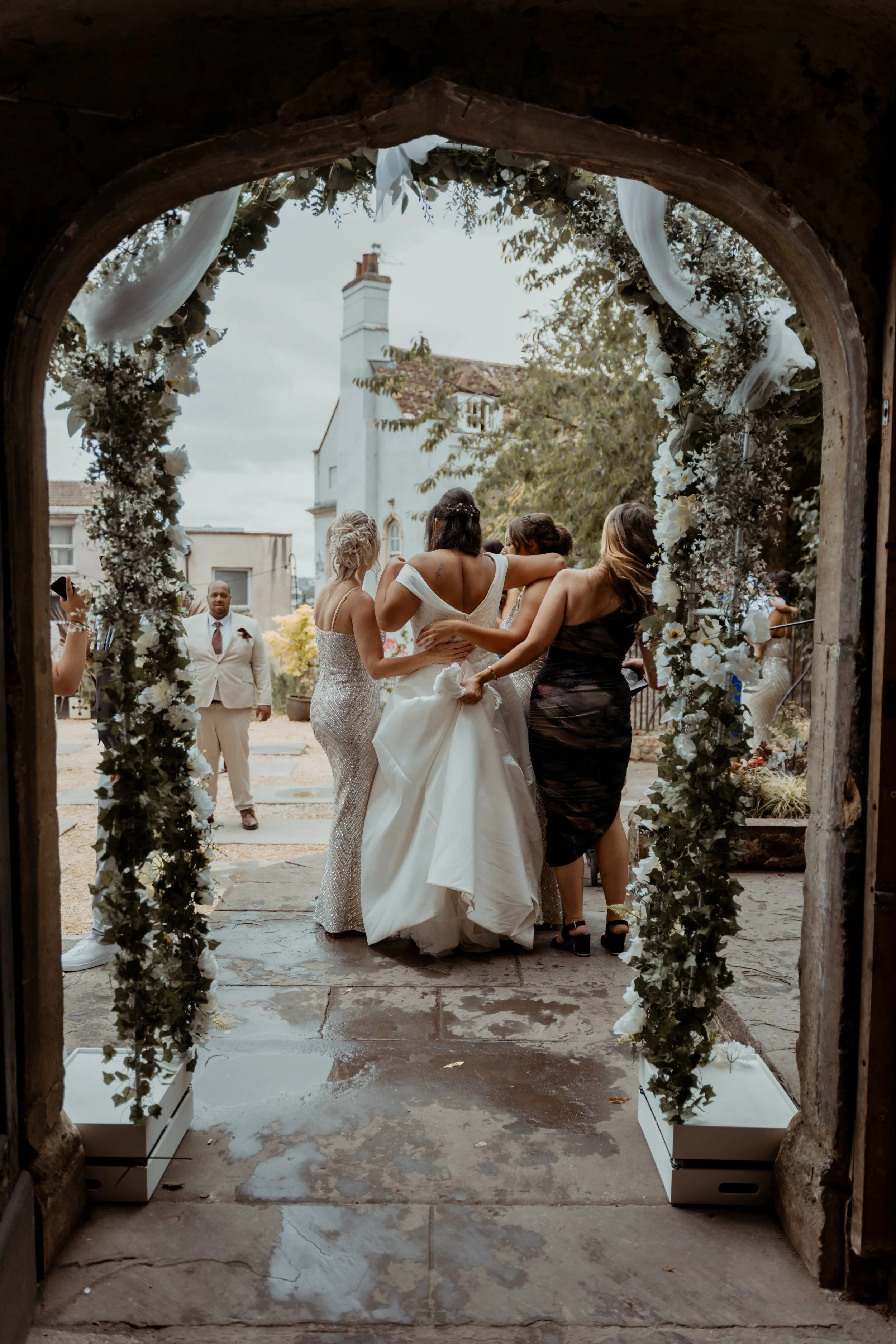 A bride being greeted by friends and family outside a decorated archway at her wedding.