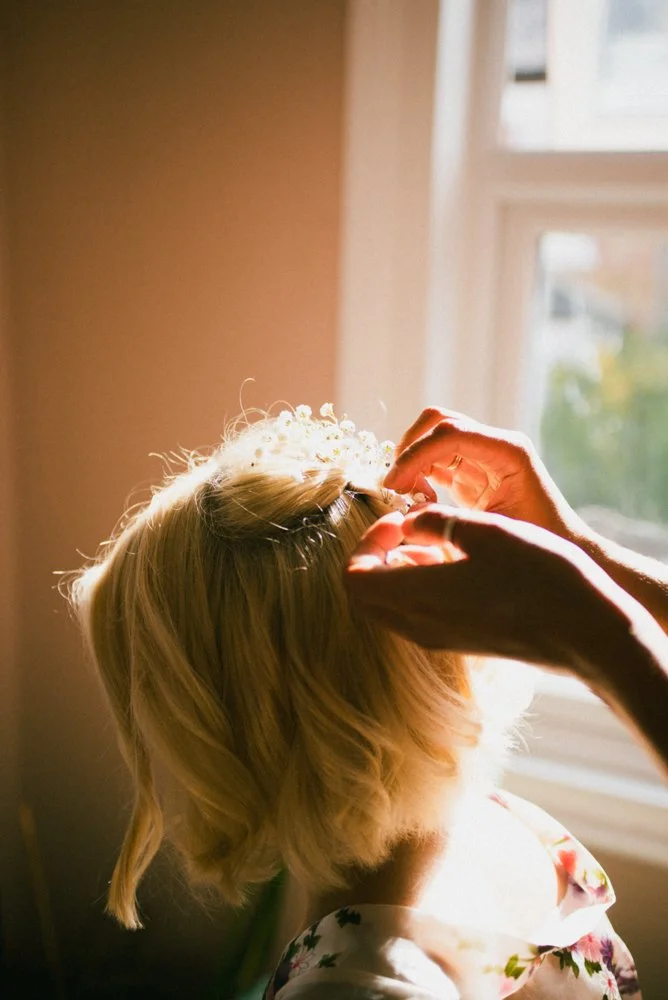 A woman with blonde hair getting a tiara placed on her head by another person in a sunlit room.