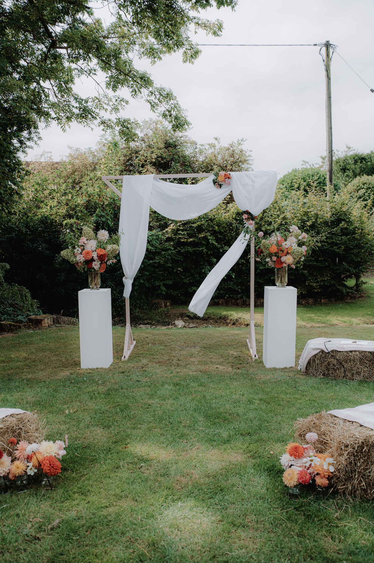 Outdoor wedding altar with white drapery and floral arrangements, set on grassy lawn with hay bales and trees in the background.