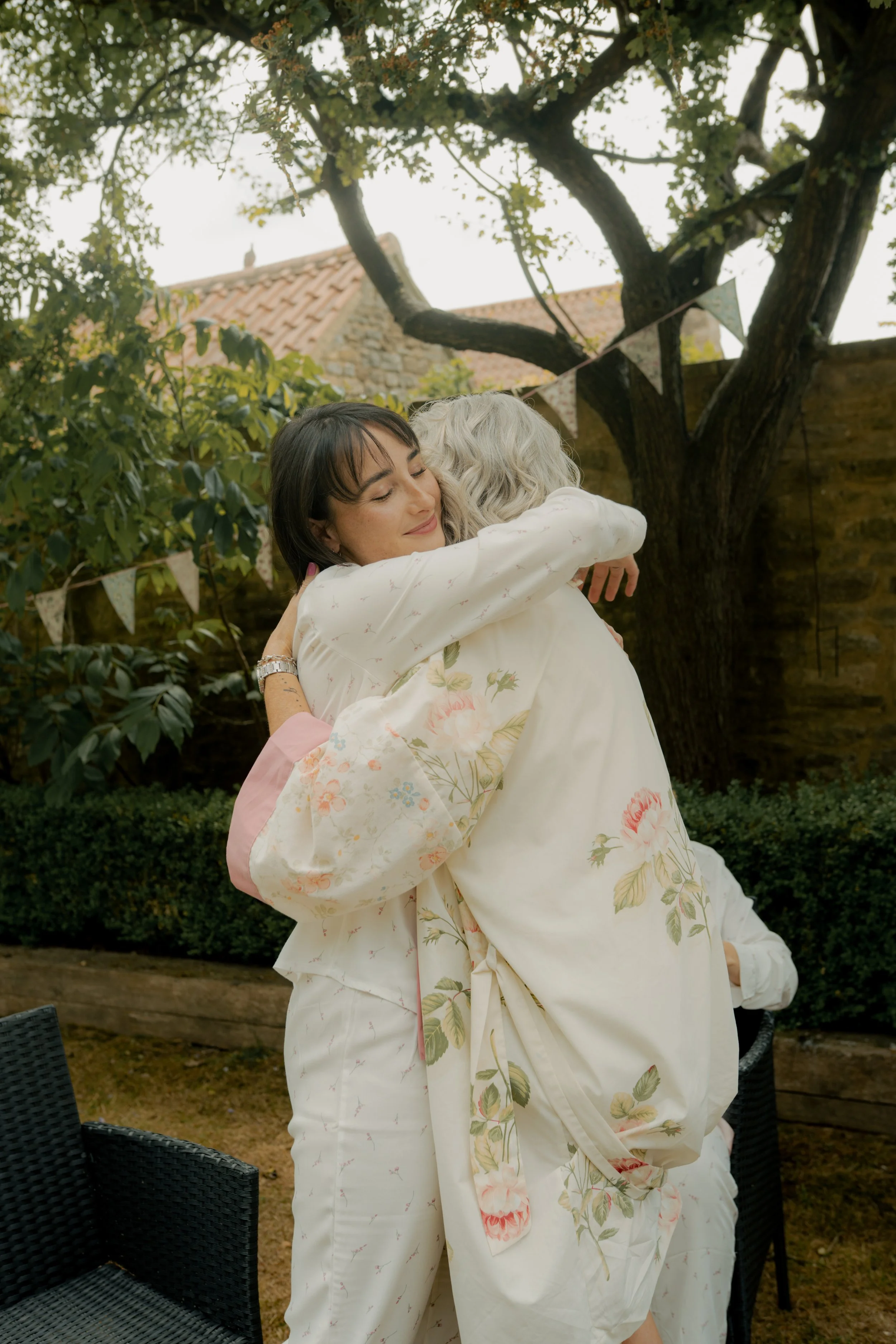 Two women hugging outdoors in a garden with a stone wall, a large tree, and bunting banners hanging between branches.