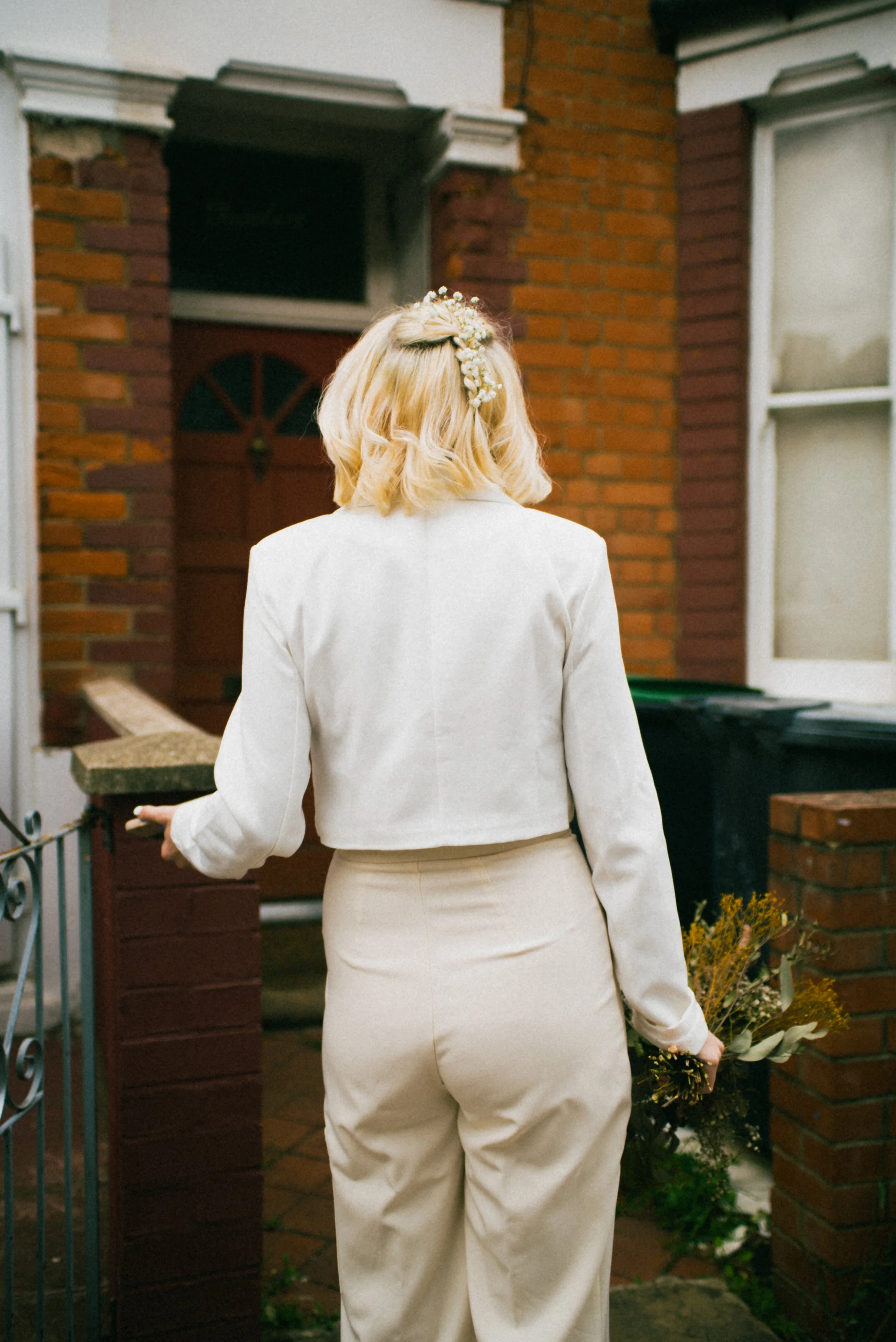 A woman with blonde hair decorated with a floral hairpiece, wearing a white jacket and cream-colored pants, is standing outside in front of a brick house, holding a bouquet of dried flowers.