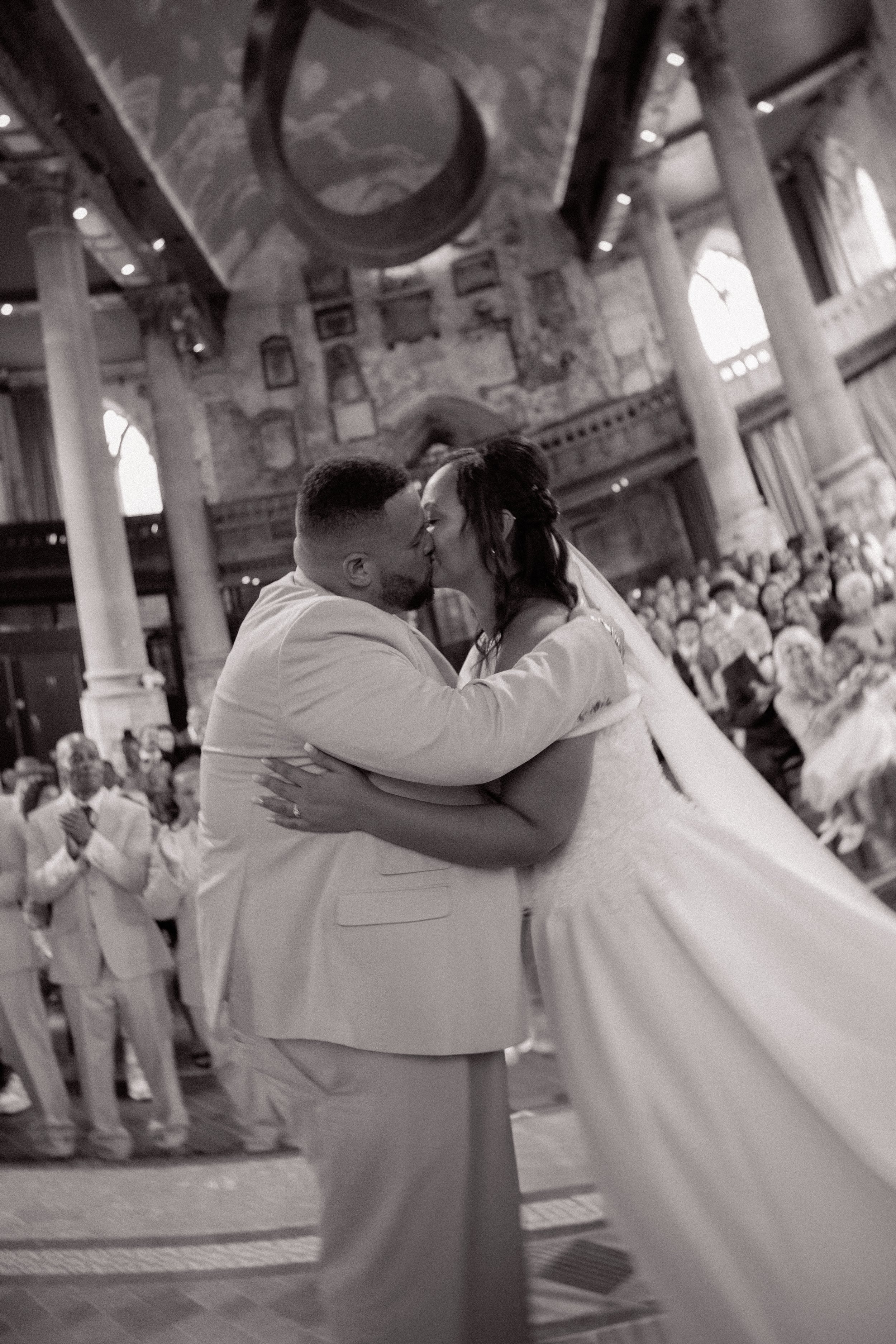 A wedding couple sharing a kiss during their ceremony inside a historic church with high ceilings and large stained glass windows.