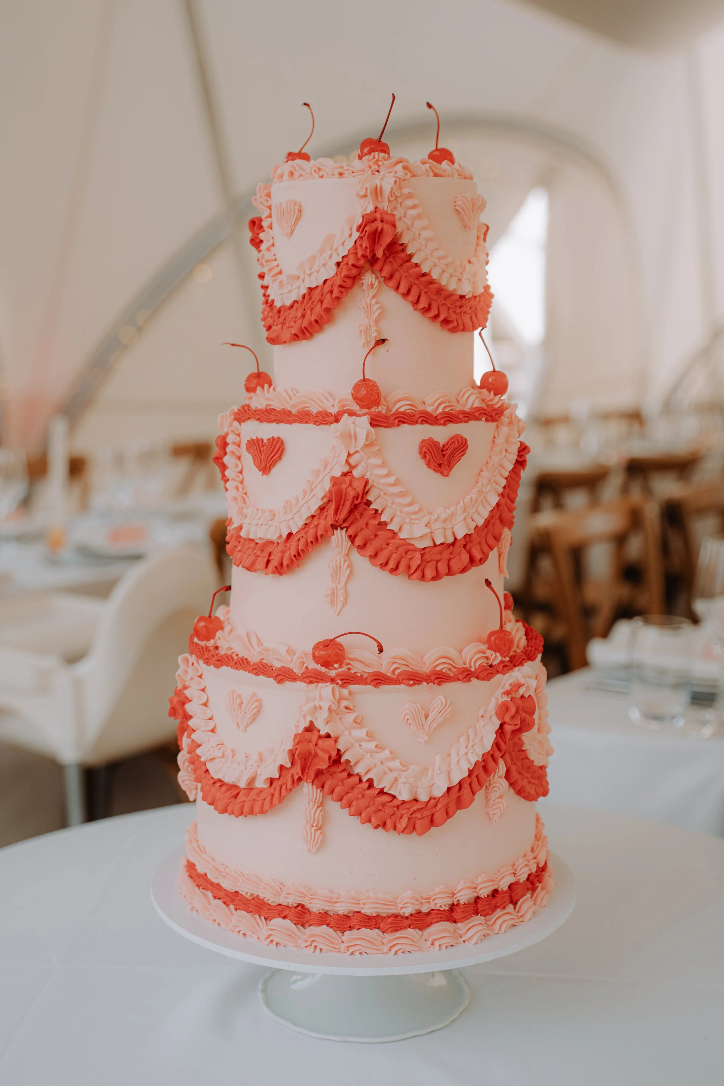 Three-tiered wedding cake decorated with pink and red icing, cherries, and heart-shaped embellishments, displayed on a white cake stand in a decorated reception hall.