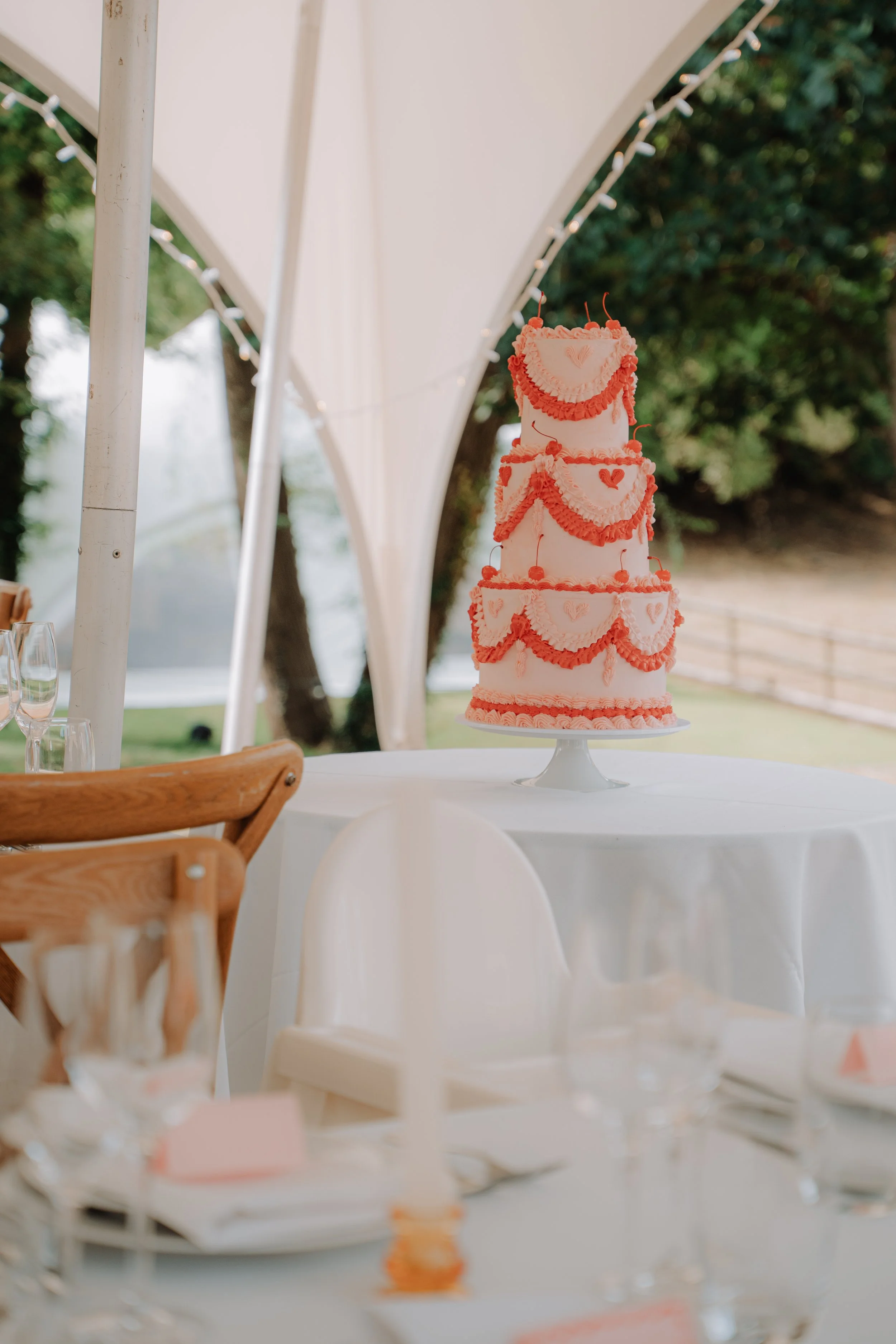 A pink and white tiered wedding cake decorated with pink icing hearts and red garlands, sitting on a white cake stand on a table at an outdoor celebration.