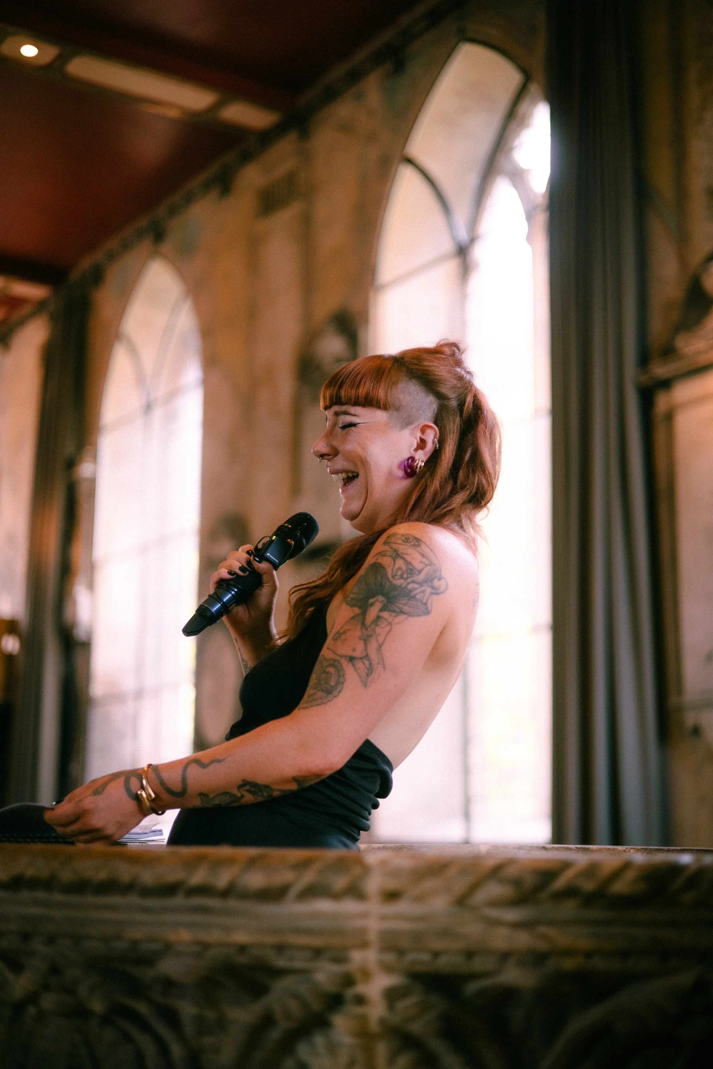 A woman with tattoos and red hair, wearing a black tank top, laughing while holding a microphone in a room with large arched windows and high ceilings.