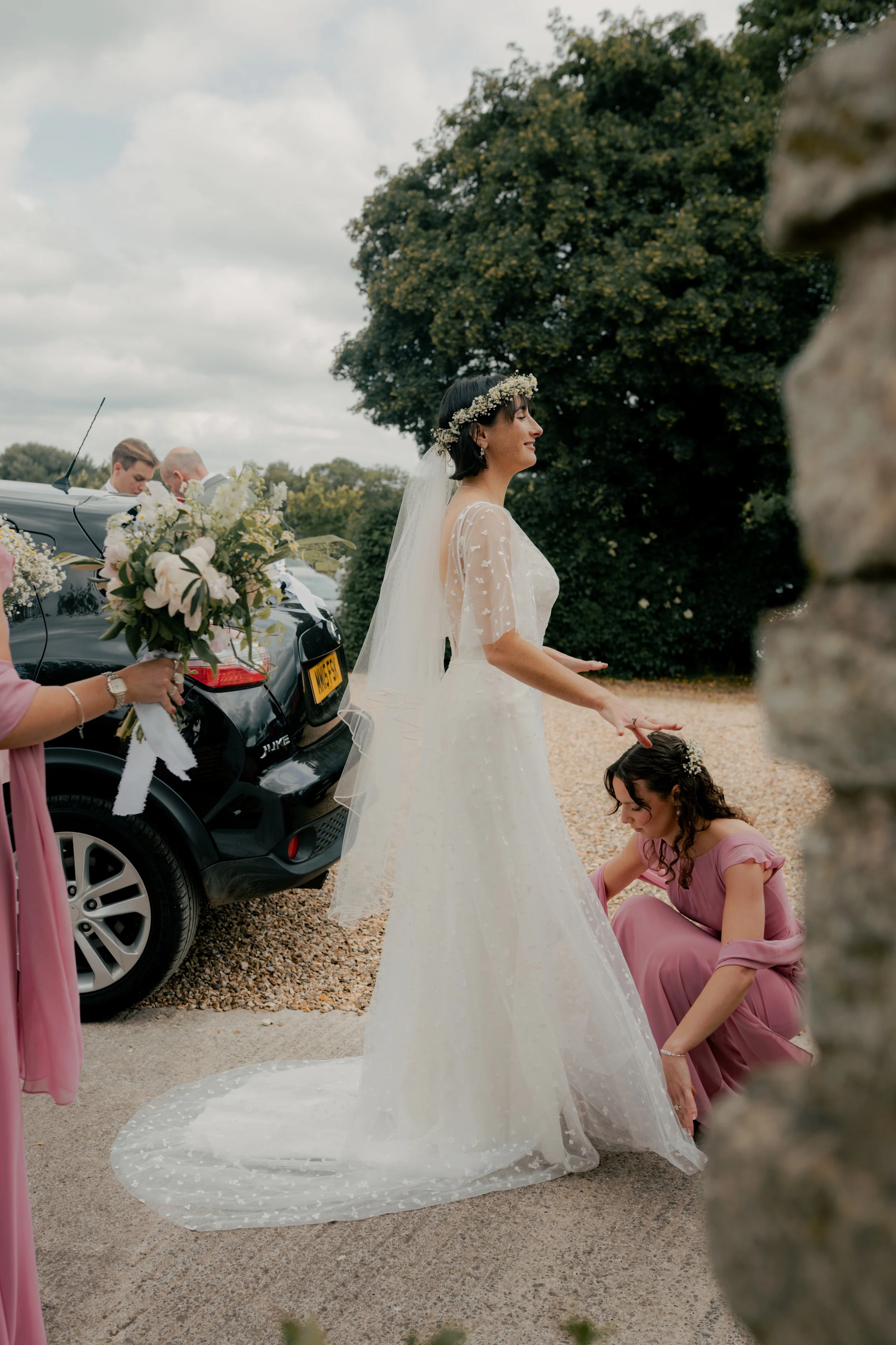 A bride in a white wedding dress with a veil and floral crown stands outside on a gravel surface, with a bridesmaid in a pink dress kneeling beside her adjusting her dress. A woman holding a bouquet is partially visible on the left, and a black car a