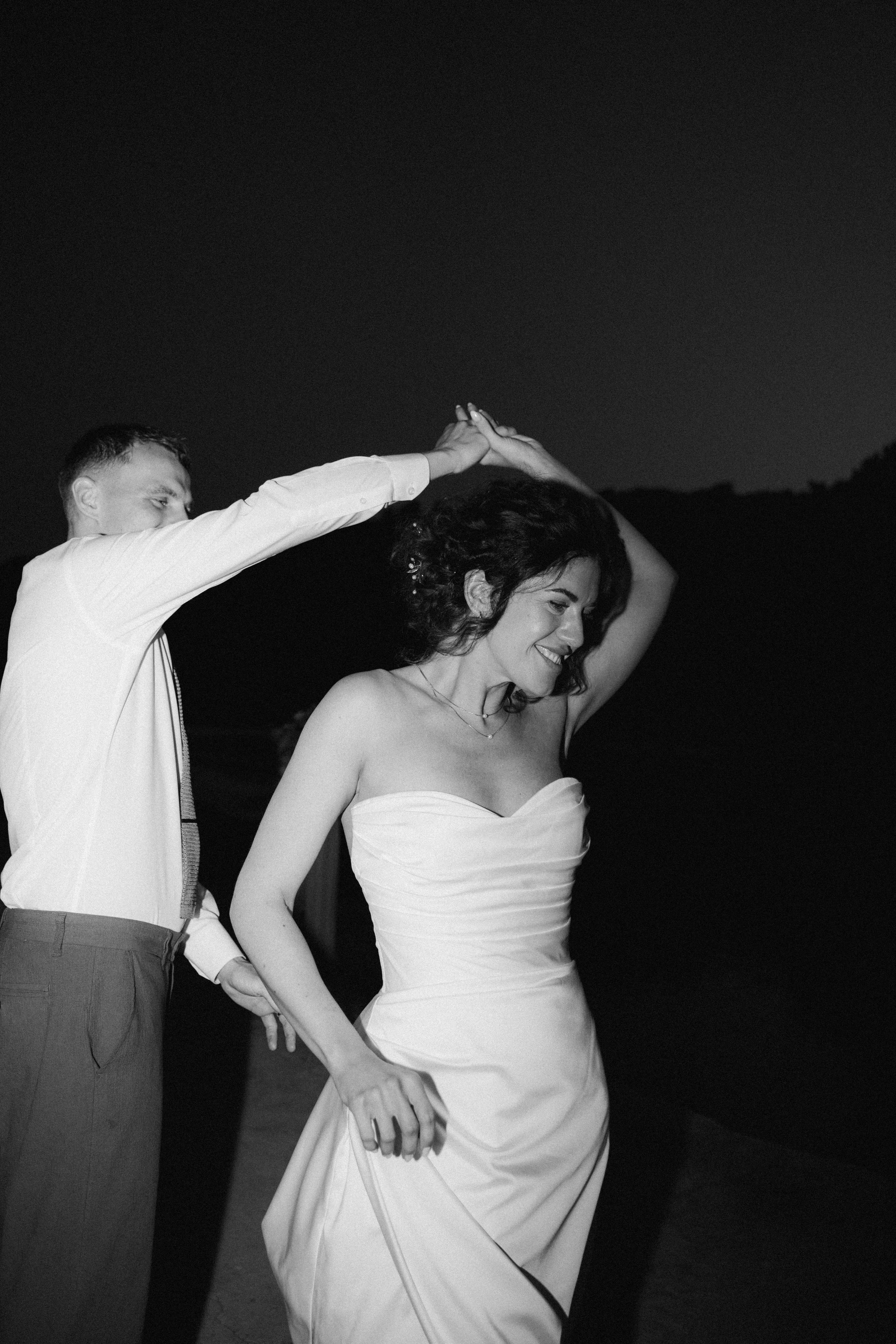 A couple dancing outdoors at night, the woman wearing a strapless dress and the man in a white shirt and suspenders, holding hands above her head.