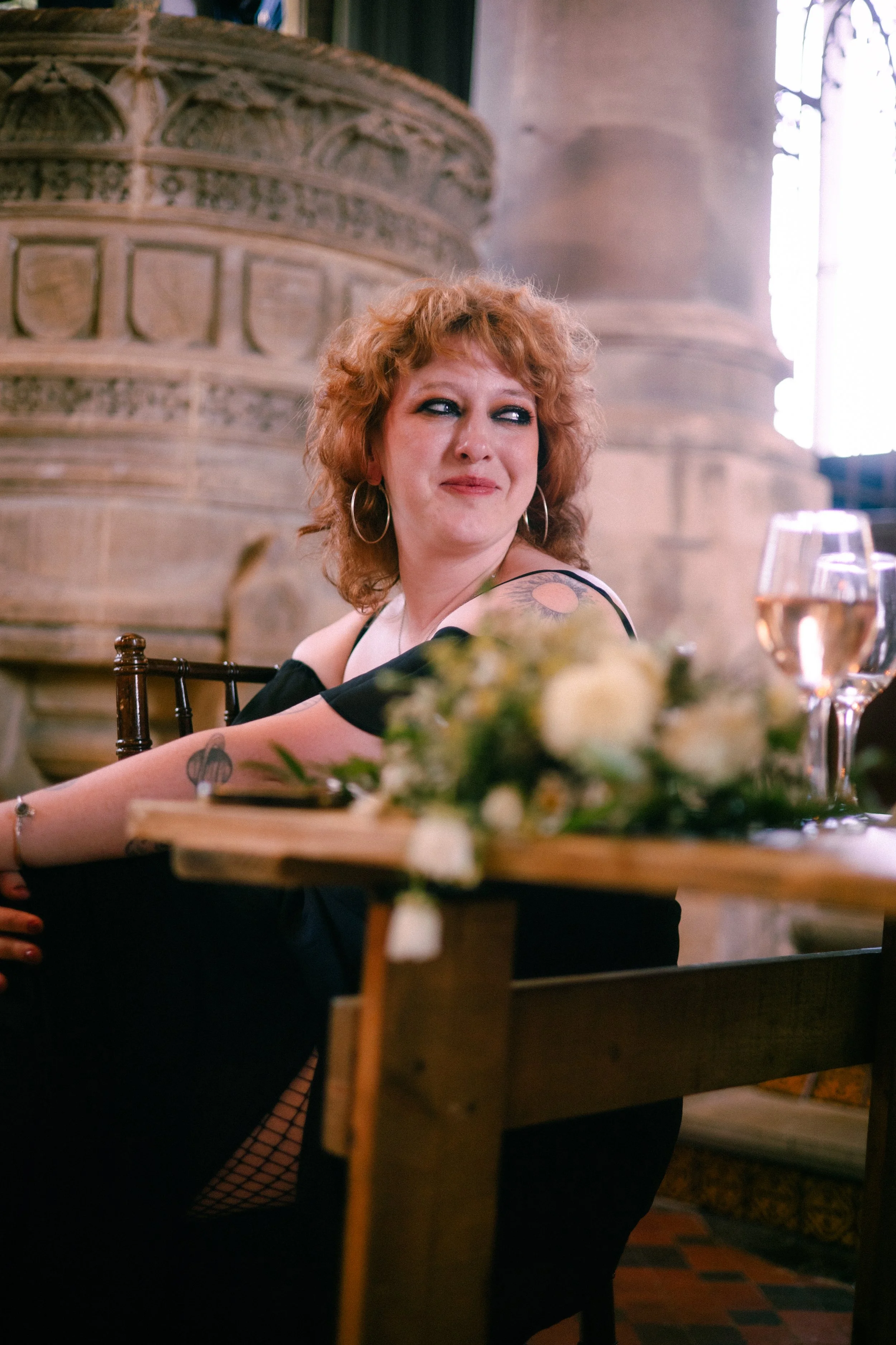 A woman with curly red hair, wearing hoop earrings and a black off-shoulder top, is seated at a table with a blurred floral centerpiece and glass of rosé wine. She is smiling and looking to her right, in a dimly lit room with ornate stone architectur