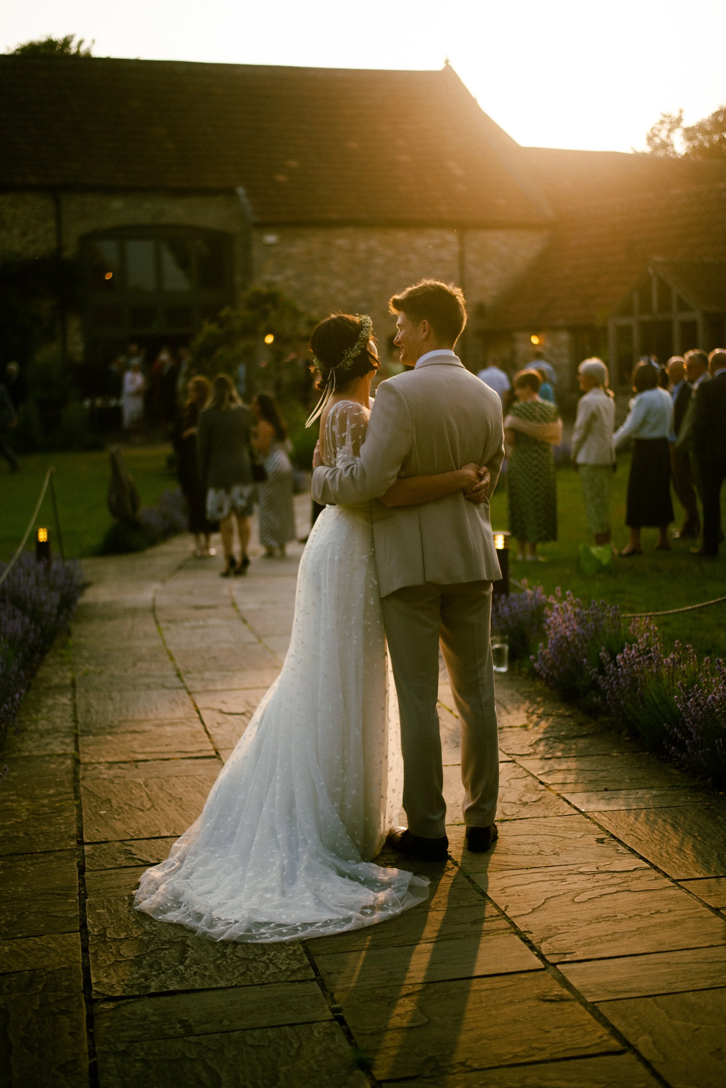 A bride and groom share a dance outdoors during their wedding at sunset, with guests in the background and a stone pathway leading to a rustic building.