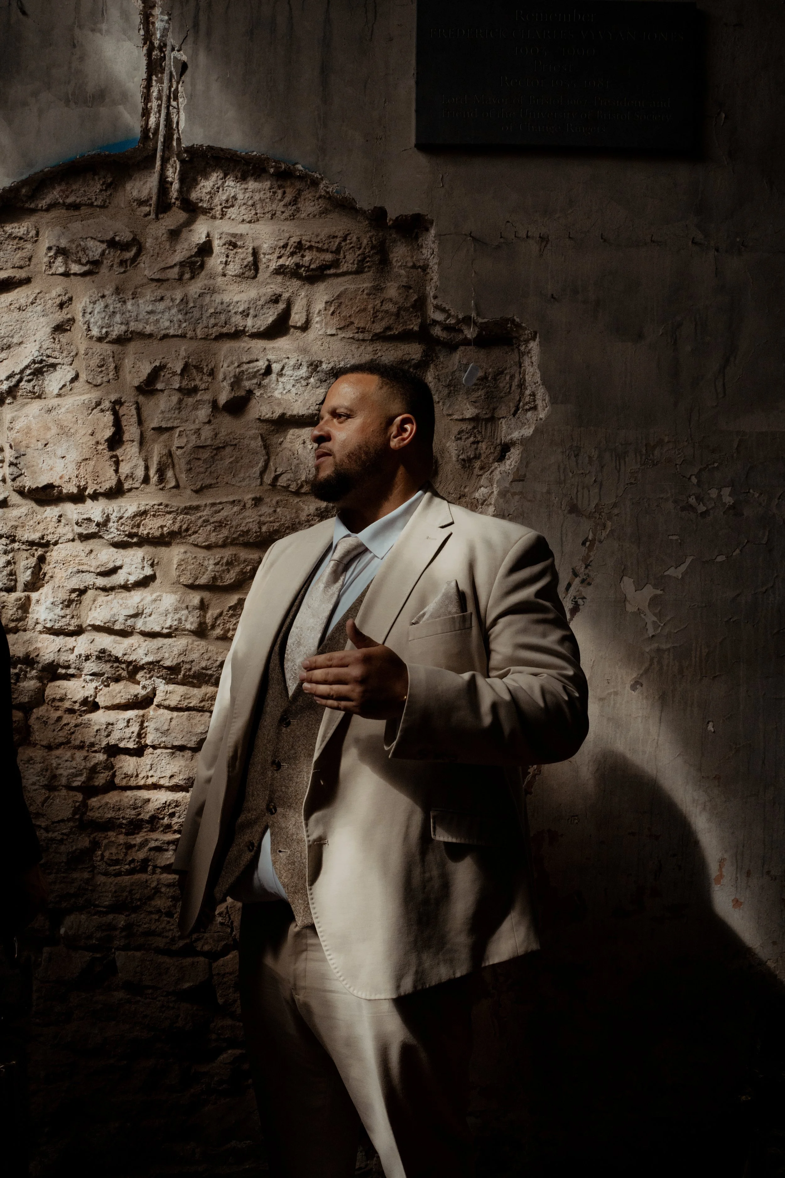 Man in a beige suit with a vest and tie, standing against a brick wall with peeling paint, with his hand over his heart, appearing to be in a moment of reflection or solemnity.