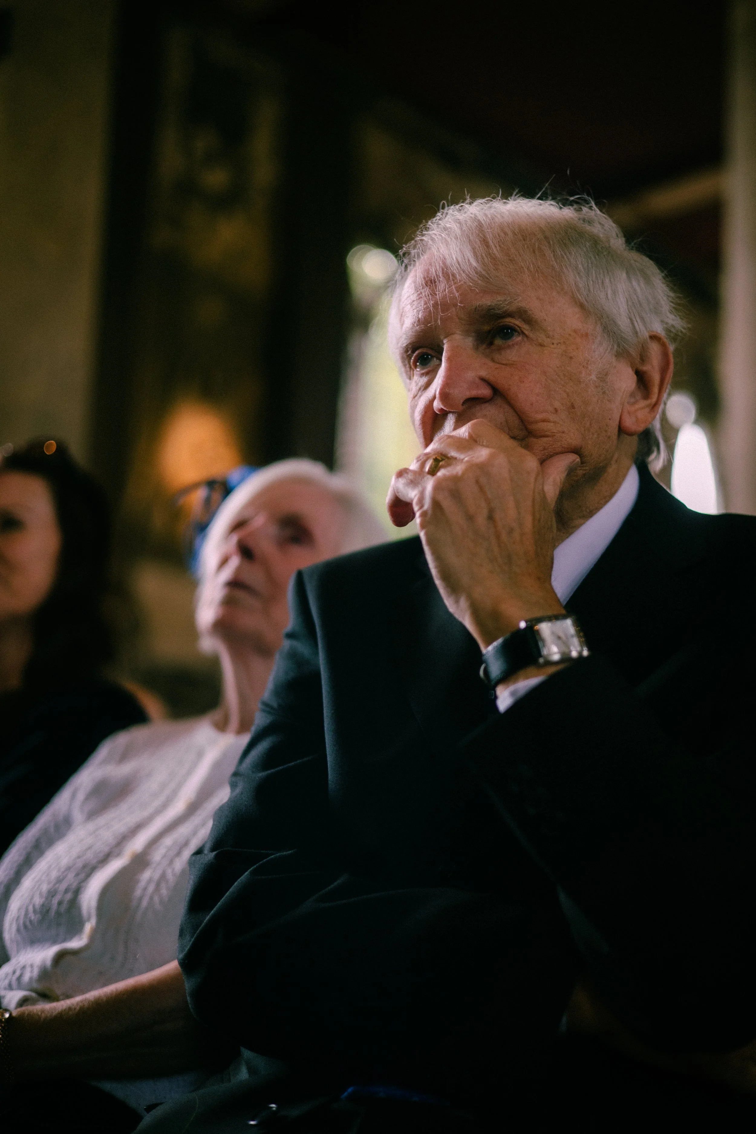 An elderly man with white hair and a thoughtful expression, seated indoors with other people, wearing a dark suit, white shirt, and a watch, with a woman in a grey sweater sitting nearby.