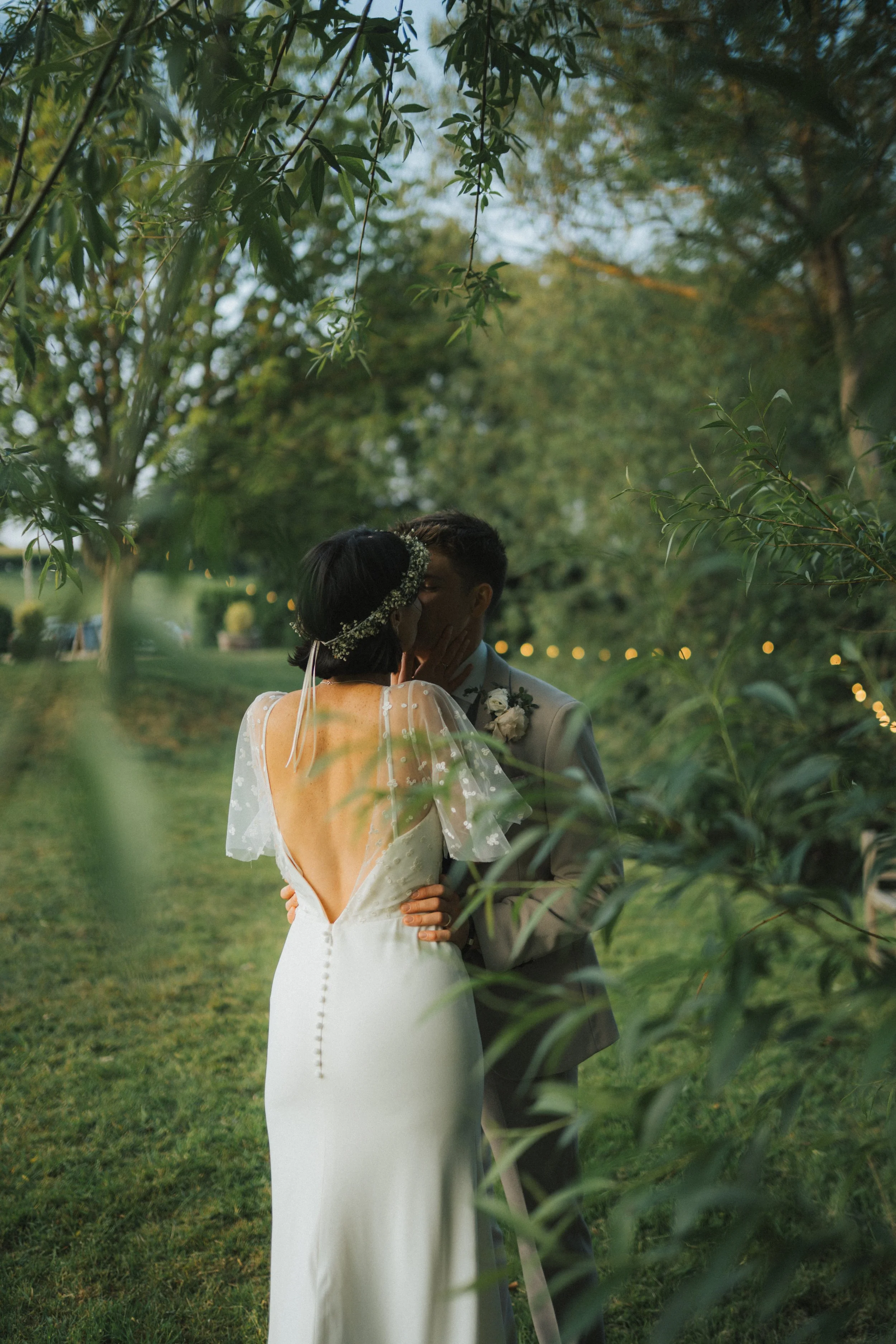 A newlywed couple sharing a kiss outdoors, surrounded by greenery and soft string lights in the background at dusk.