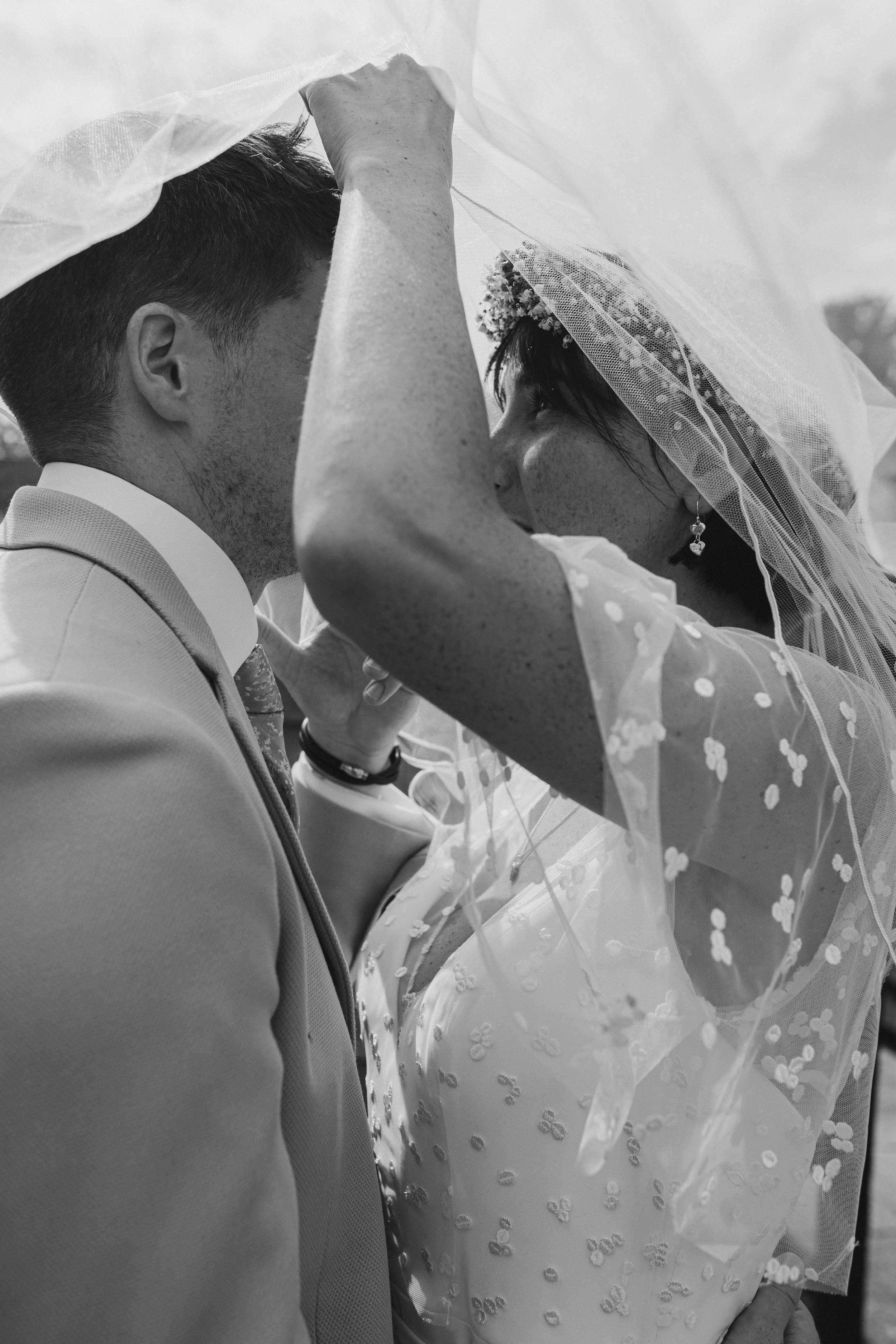 A bride and groom face each other under a veil, close-up shot in black and white.