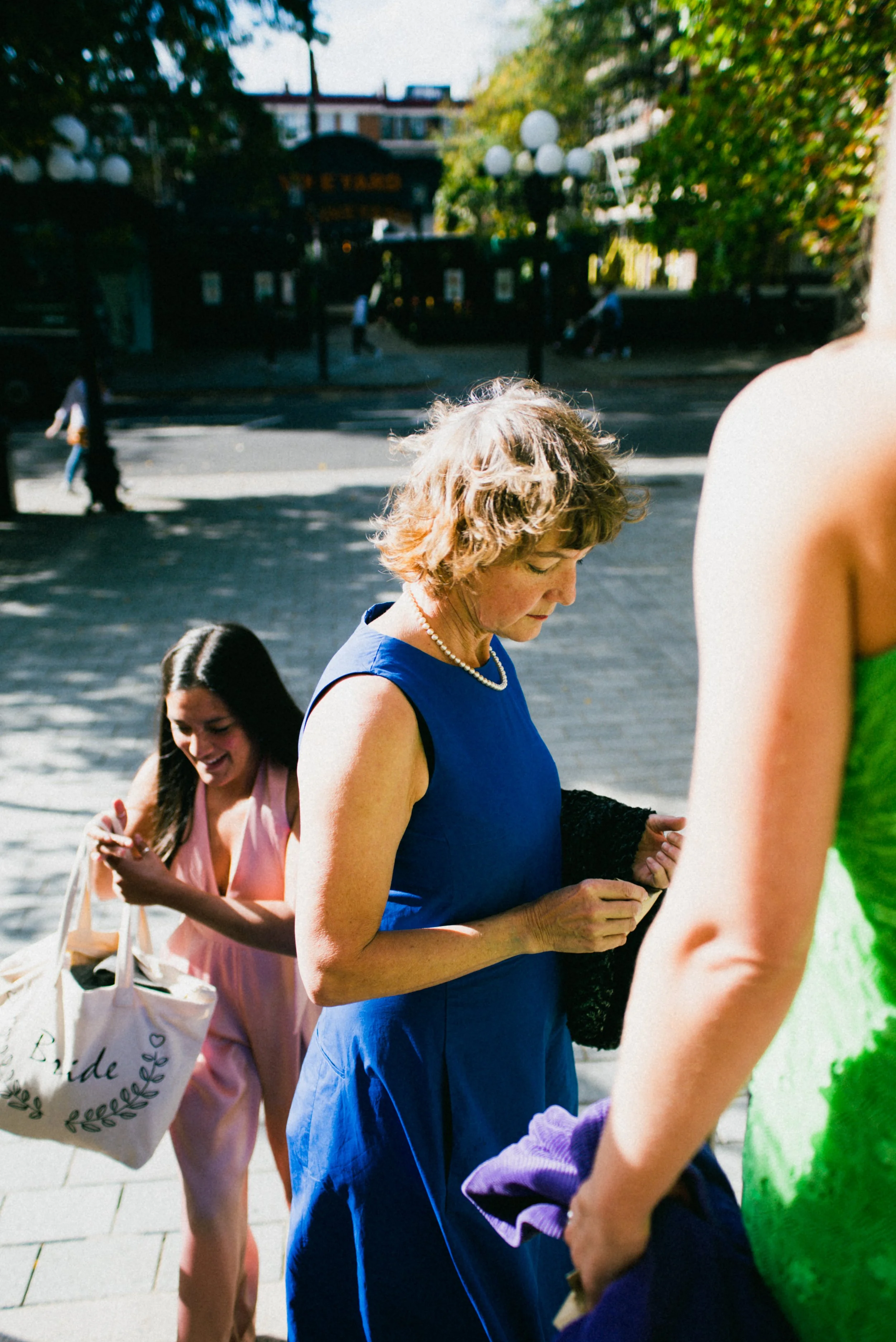 Three women preparing for a wedding outdoors on a sunny day, with trees and buildings in the background. One woman is in a blue dress, another in a pink dress with a tote bag, and the third person in a green dress is partially visible.