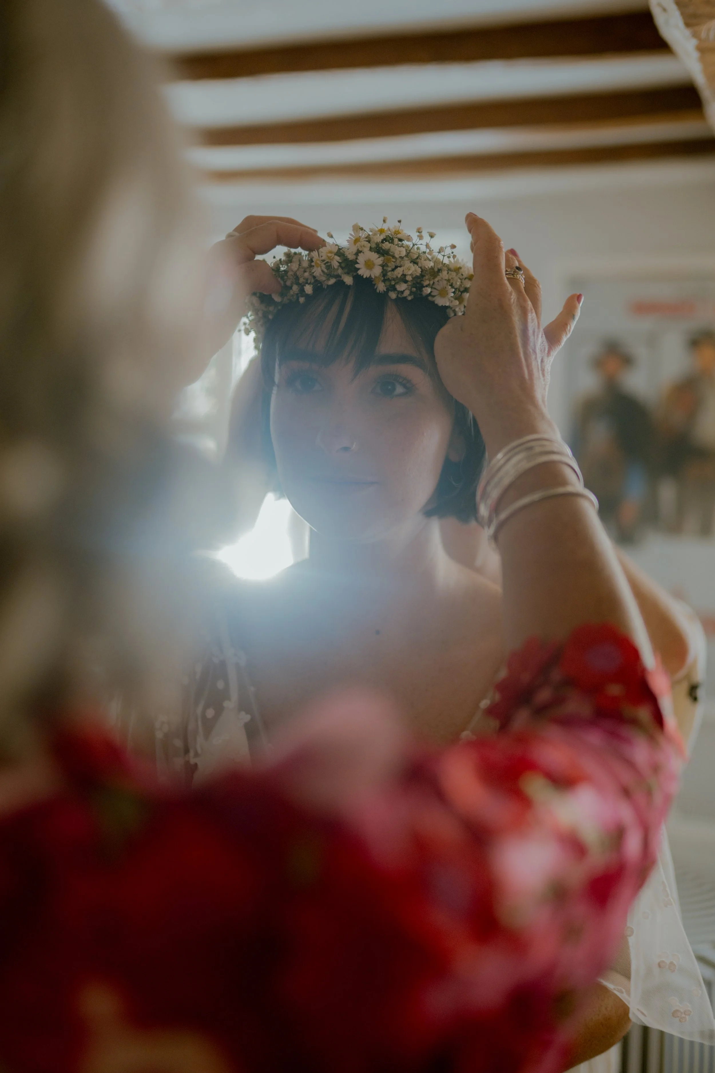 A woman wearing a floral crown is being assisted by another woman to adjust her crown, with a bright light shining behind her. The scene appears to be a moment of preparation, possibly for a wedding or special event.