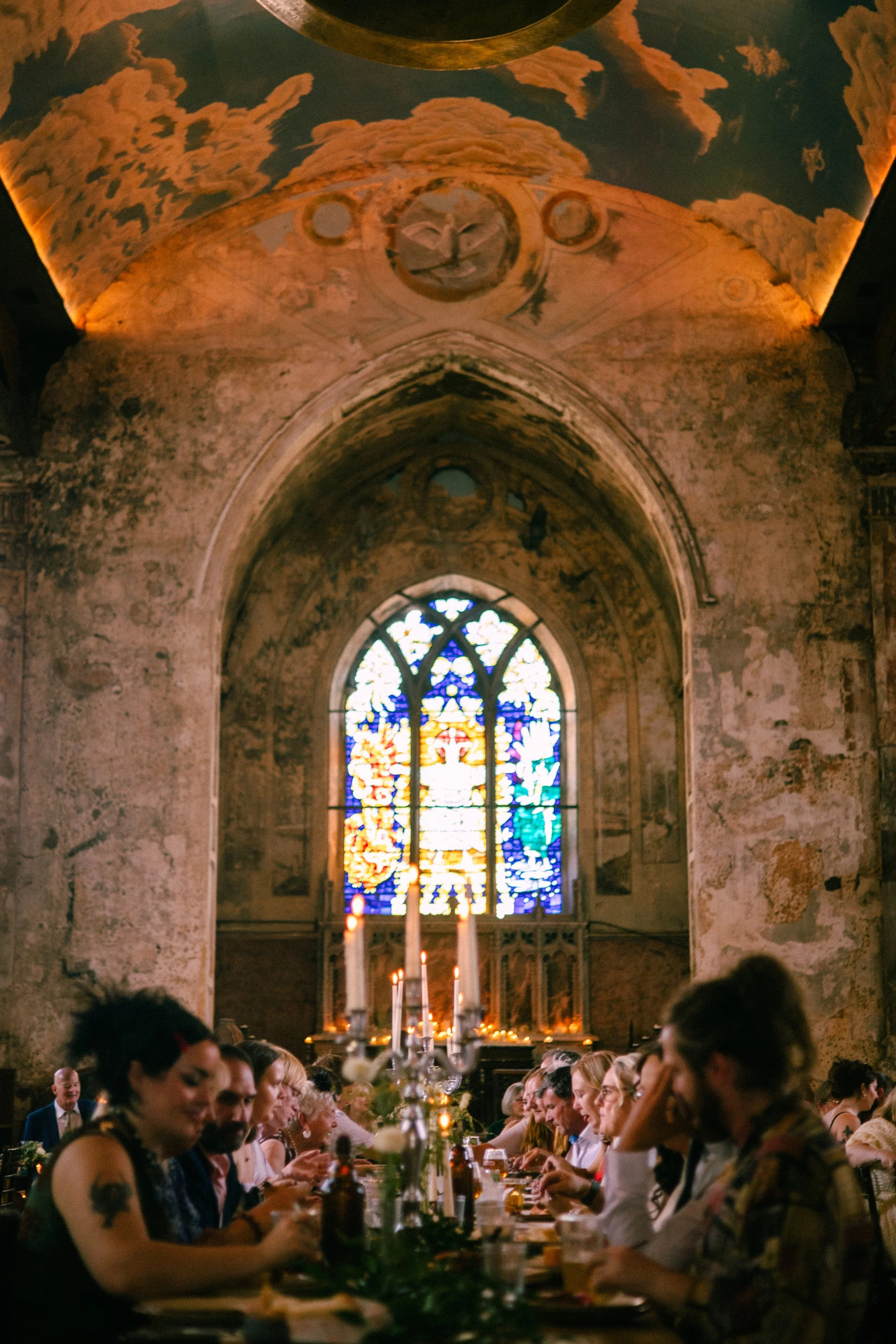 A gathering of people seated at a long table inside an old church with stained glass windows and high, arched ceilings. There are lit candles and a chandelier, creating a warm, intimate atmosphere.