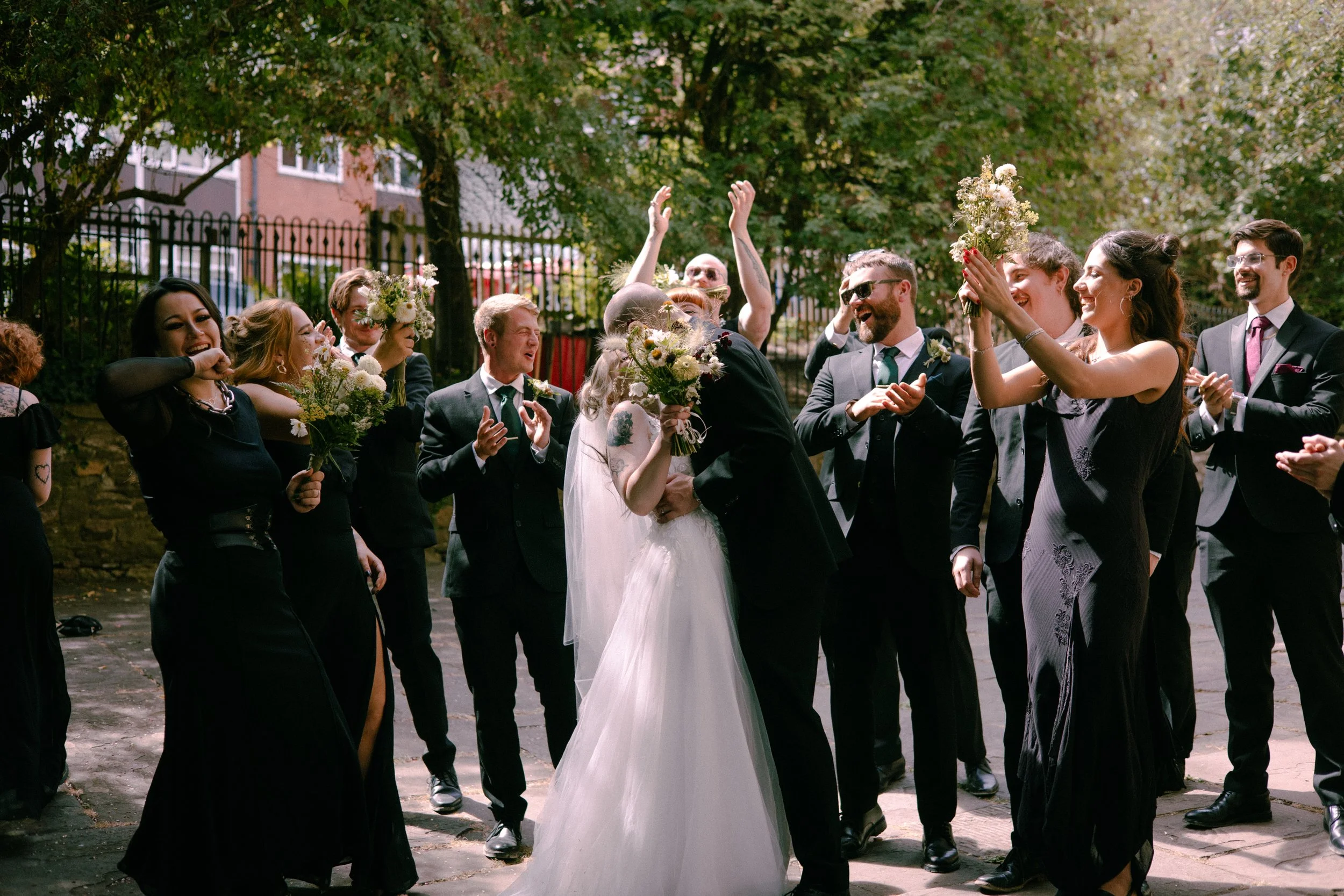 A wedding celebration outdoors with a couple kissing, surrounded by friends holding flowers and smiling.