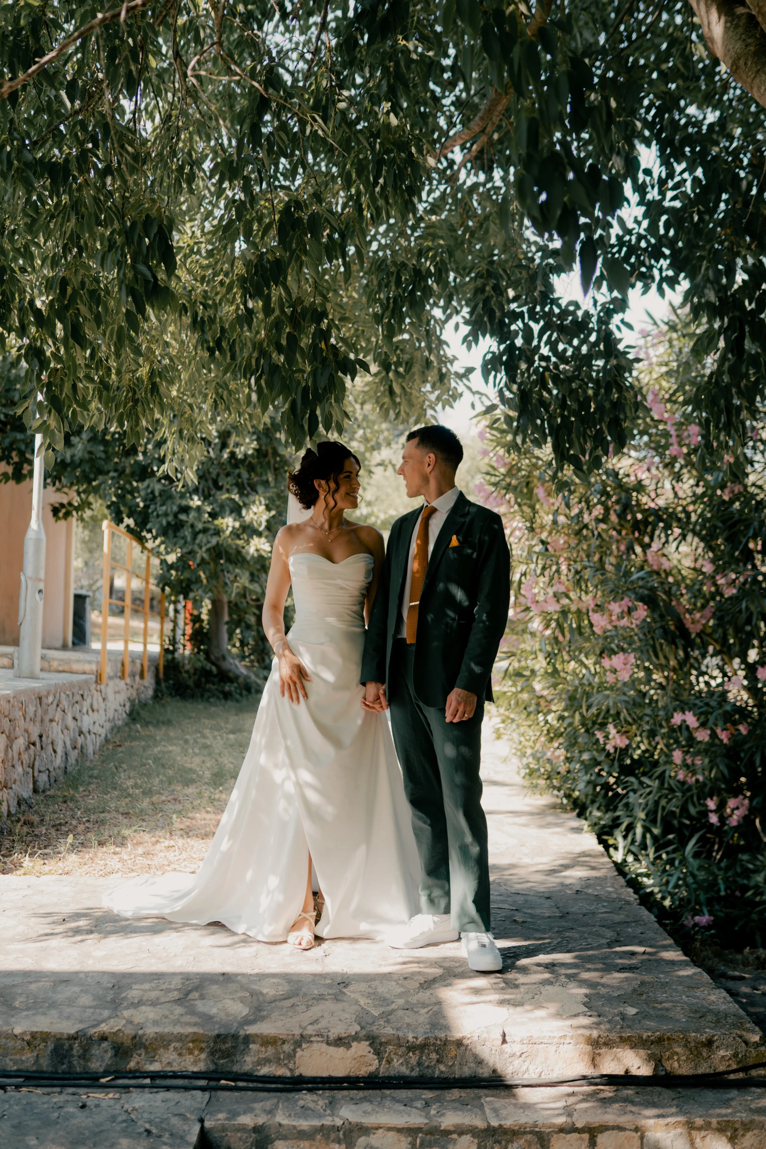 A bride and groom holding hands, standing under a tree on a stone pathway, gazing at each other, during their wedding celebration outdoors.