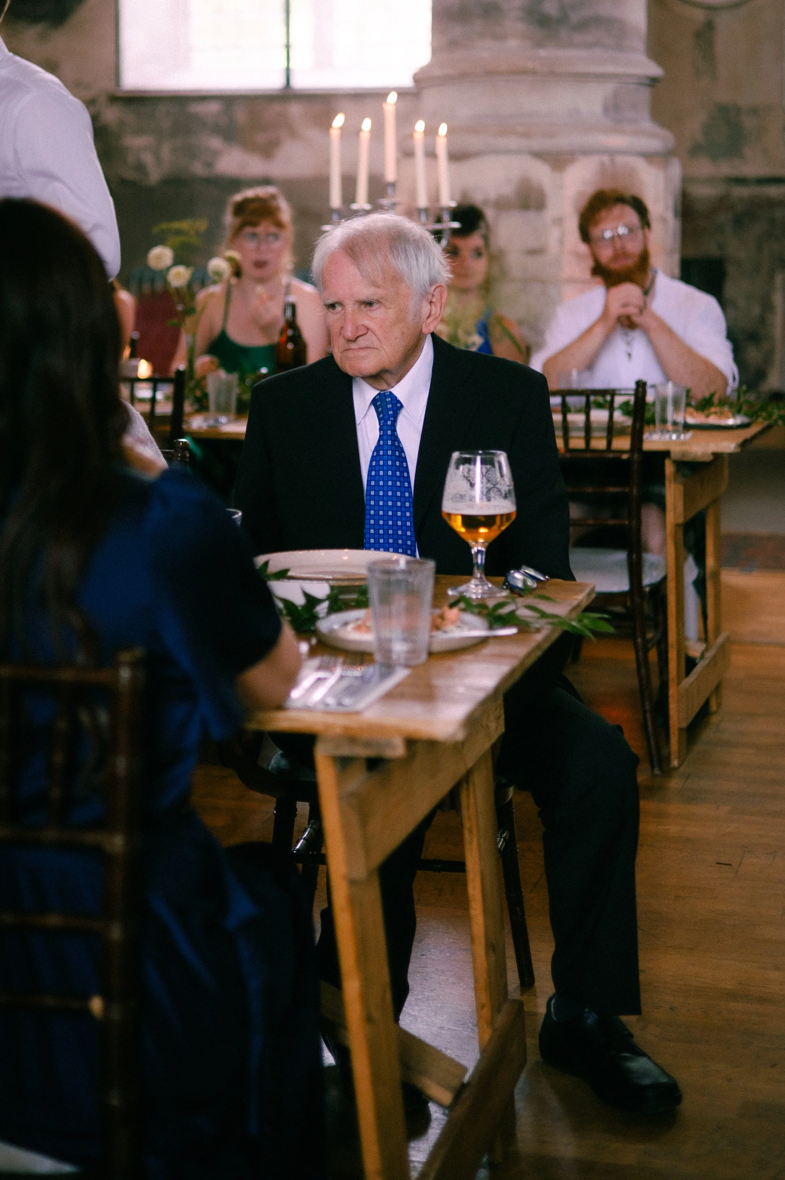 An elderly man in a black suit with a blue tie sitting at a wooden table in a restaurant, looking to the side with a serious expression. There is a glass of beer, plates, and silverware on the table. In the background, people are seated at another ta