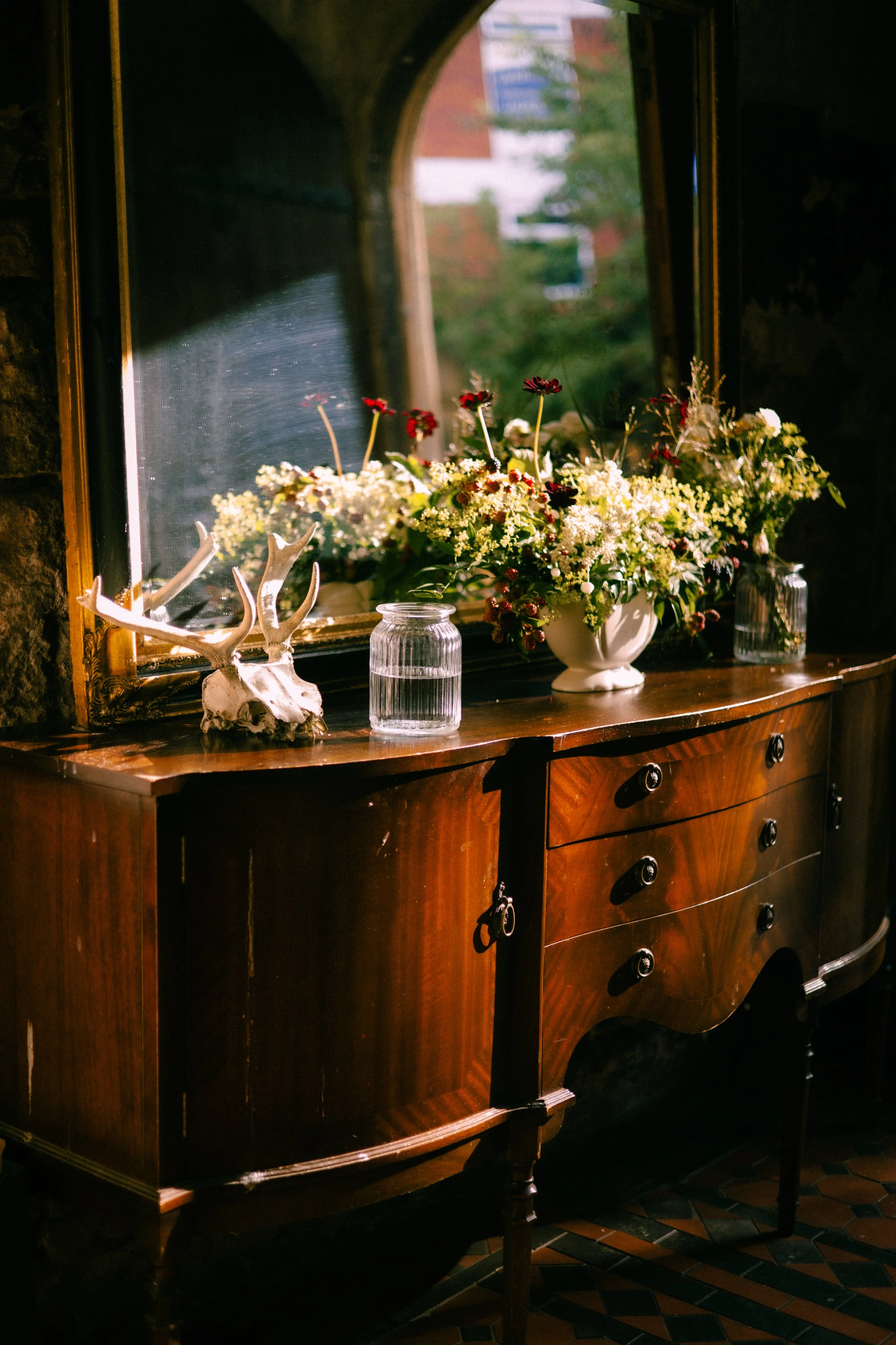 A wooden dresser with a mirror above, decorated with a flower arrangement in a white vase, a glass jar, and antlers, all placed in front of a window with a stone frame, through which a street and building are visible.