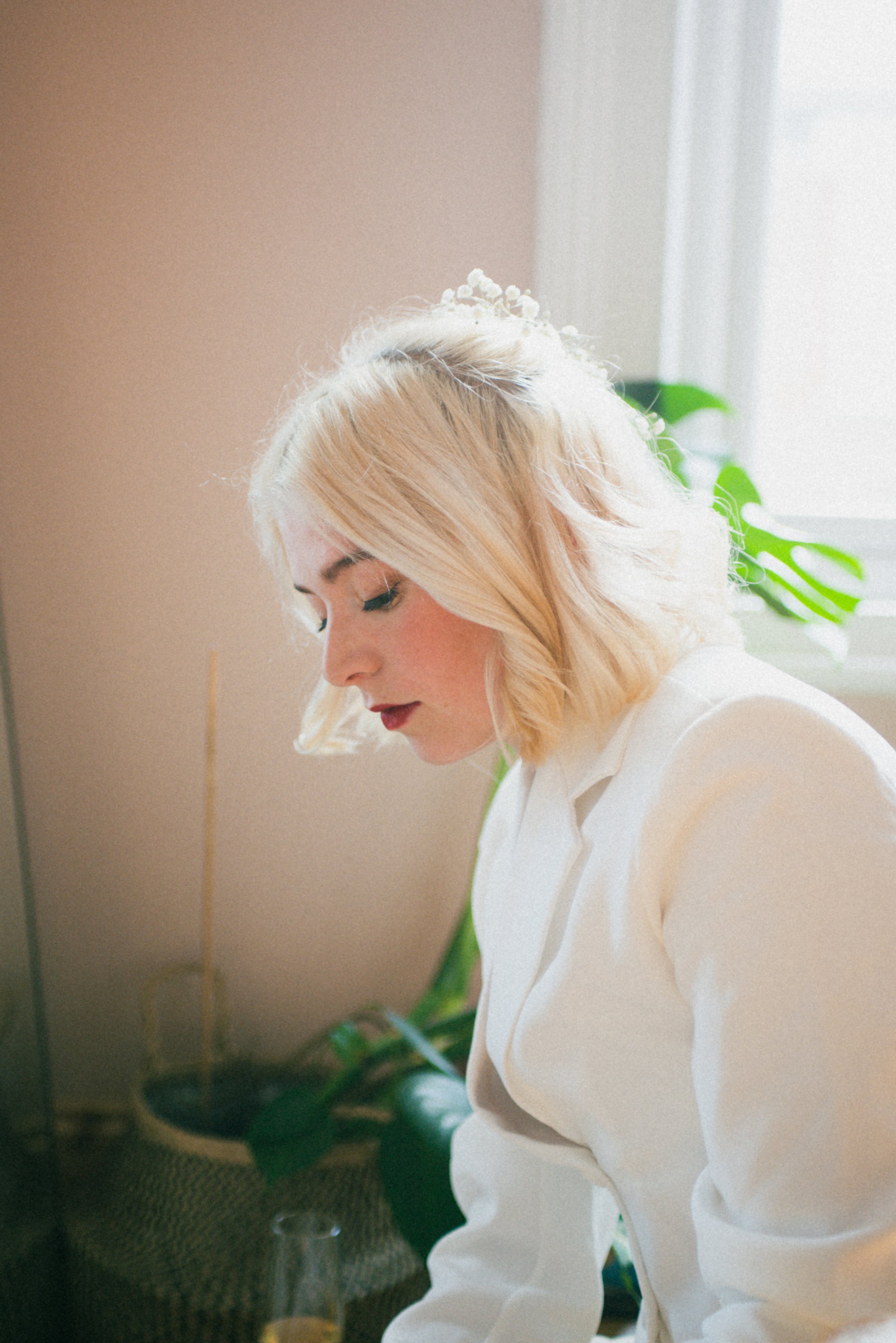 A young woman with blonde hair and red lipstick looking down, sitting indoors near a window with natural light, and a large green houseplant in the background.