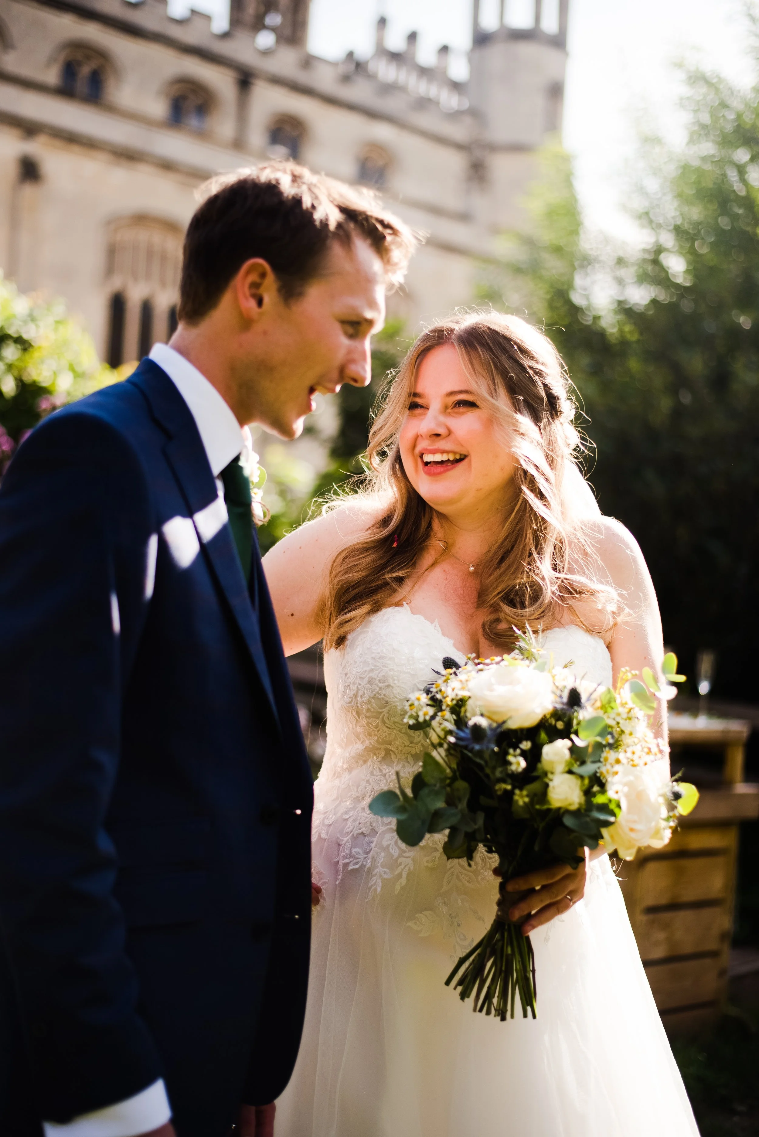 A bride and groom smiling and laughing outdoors on their wedding day, with a historic building in the background. The bride holds a bouquet of white and dark flowers.