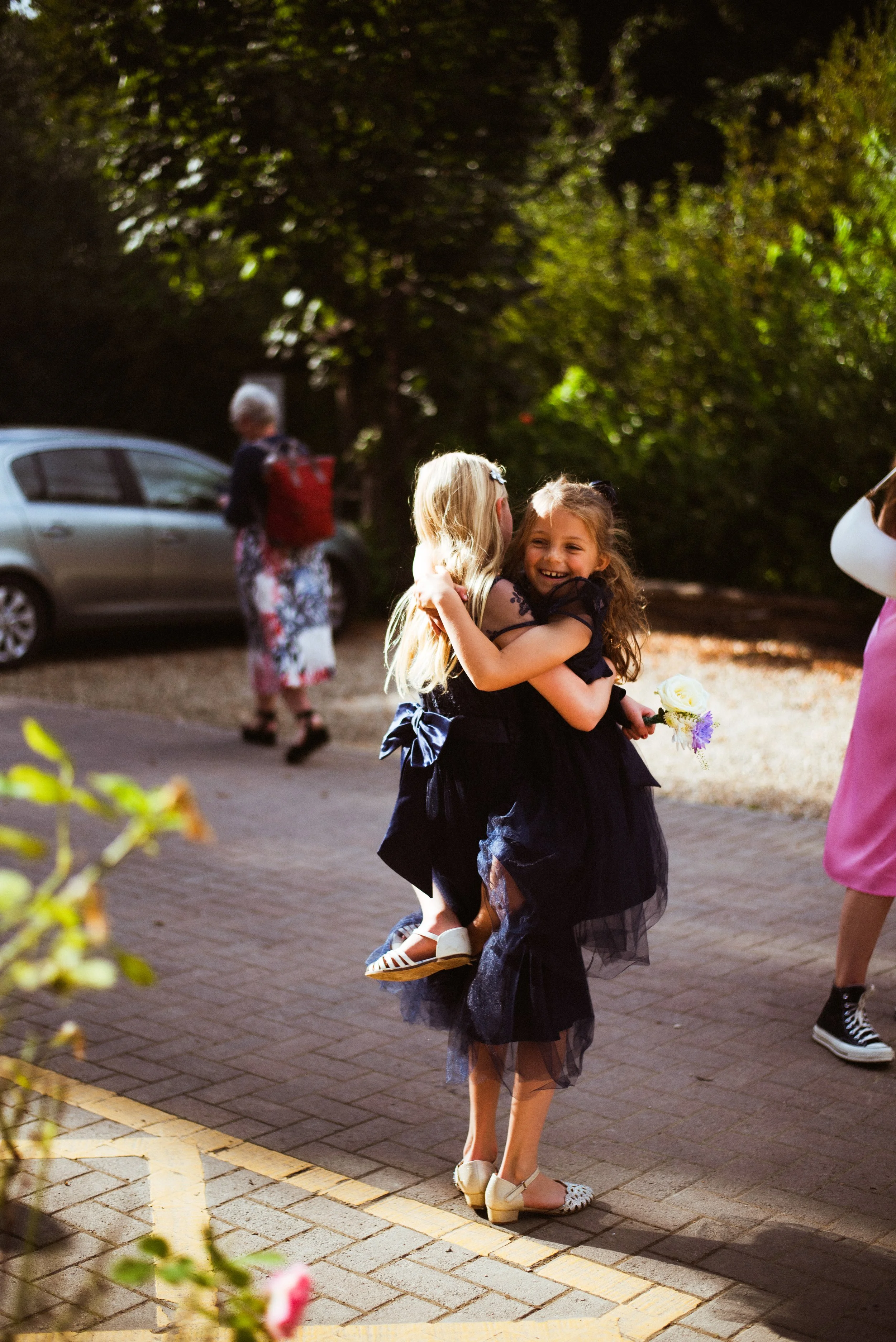 Two young girls in black dresses hugging each other in an outdoor setting, one girl holding flowers, with a car and an older woman in the background.