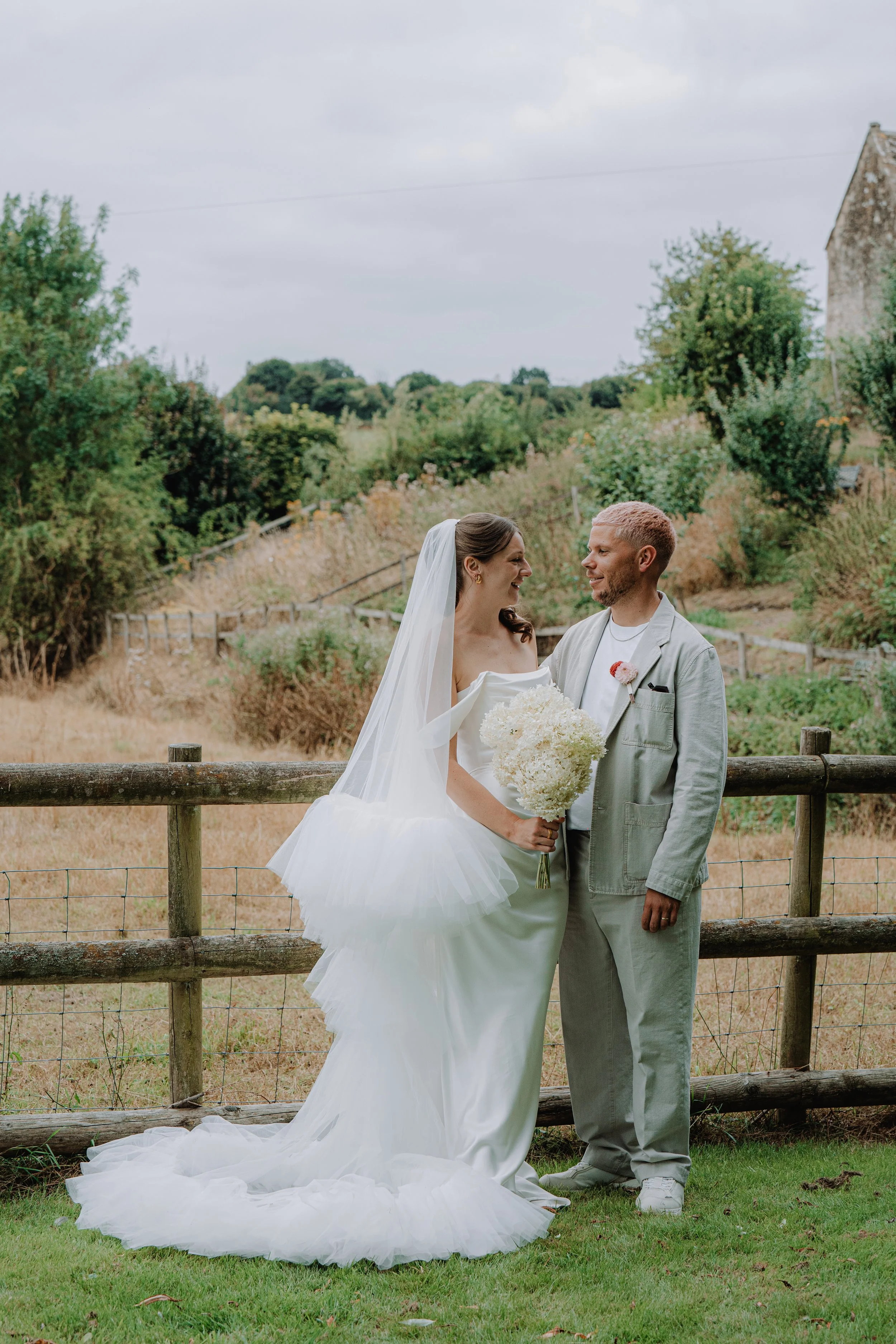 A bride and groom together outdoors, smiling and looking at each other, on a grassy area with a wooden fence and greenery in the background.