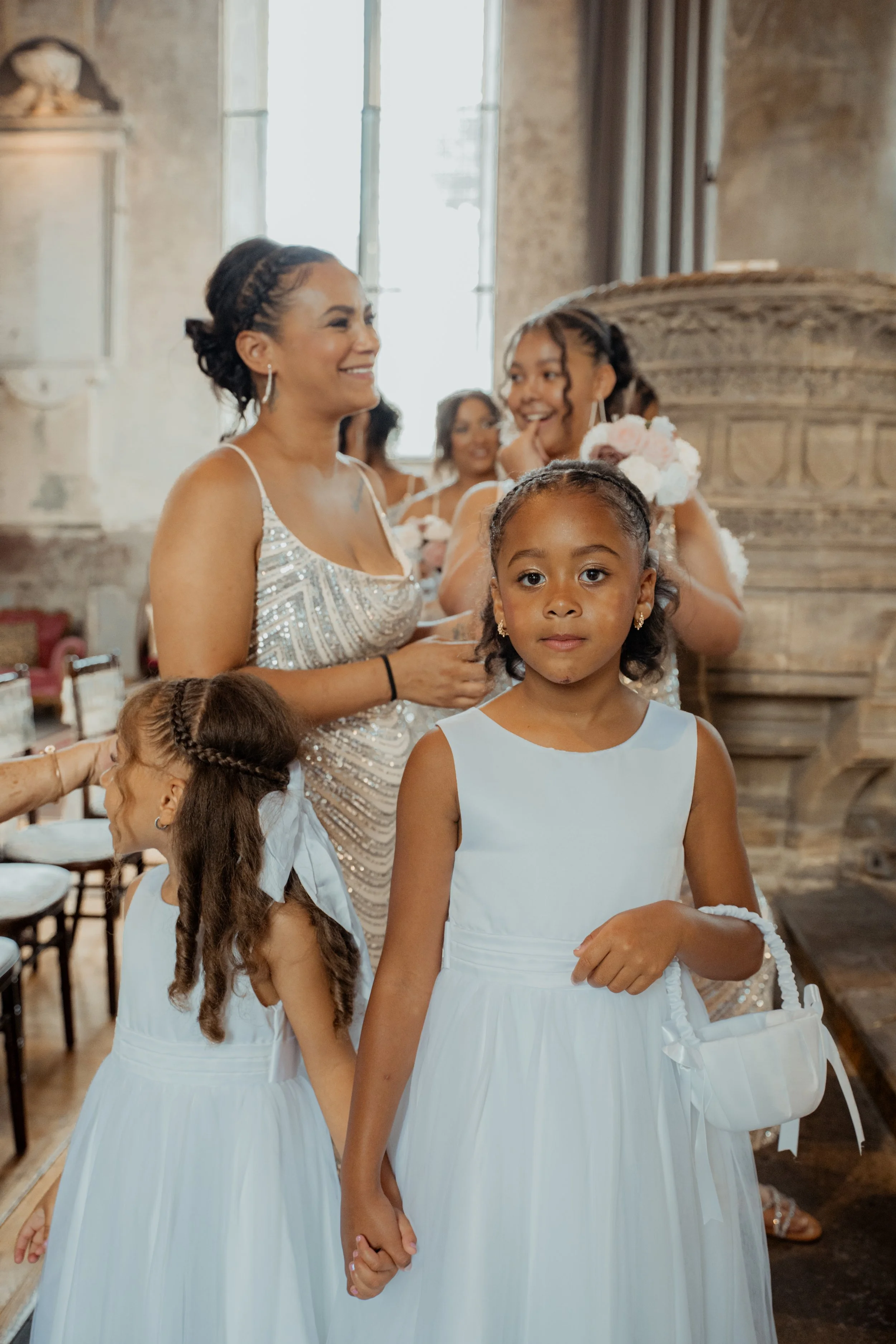 A group of women and girls dressed in white at a wedding, with the focus on a young girl holding a small basket, in an ornate indoor setting.