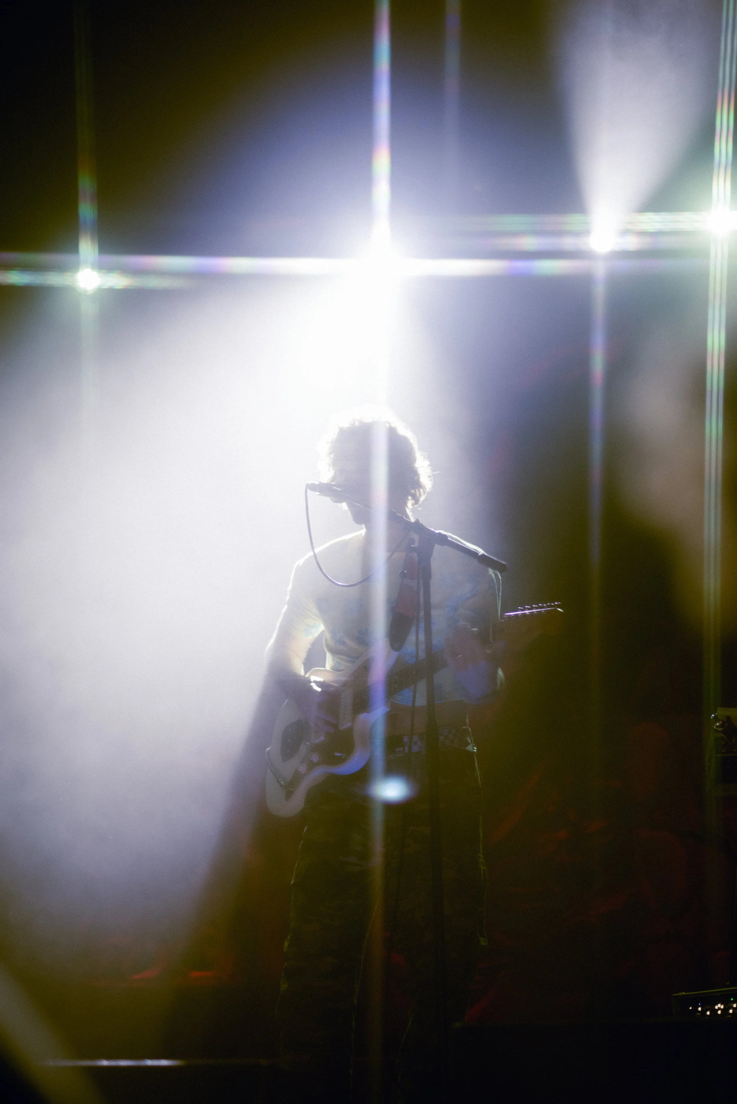 A person with curly hair playing an electric guitar on stage, illuminated by bright stage lights creating a backlit silhouette effect.