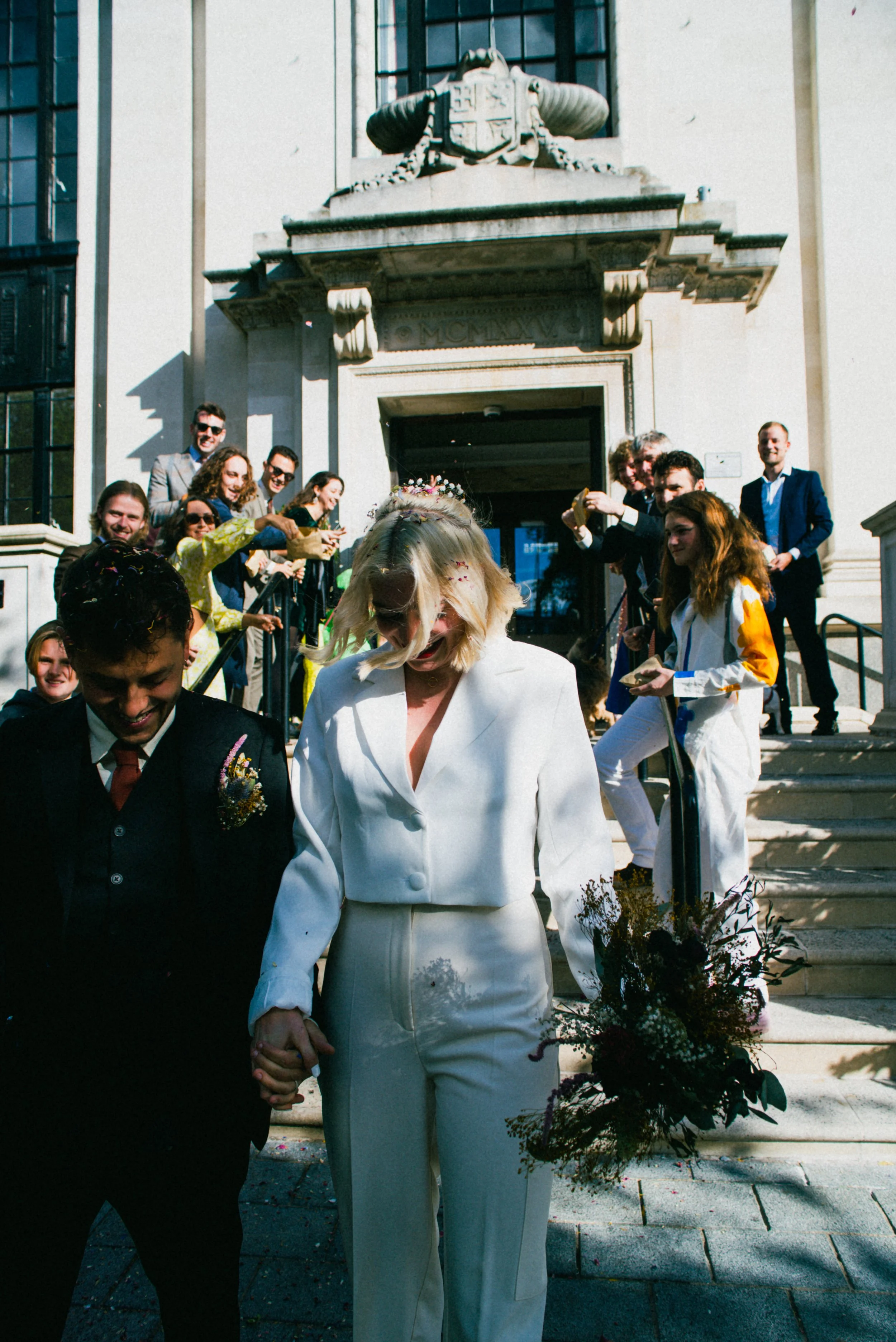 A newly married couple walks down the stairs holding hands, surrounded by celebrating friends and family, with a church building in the background.