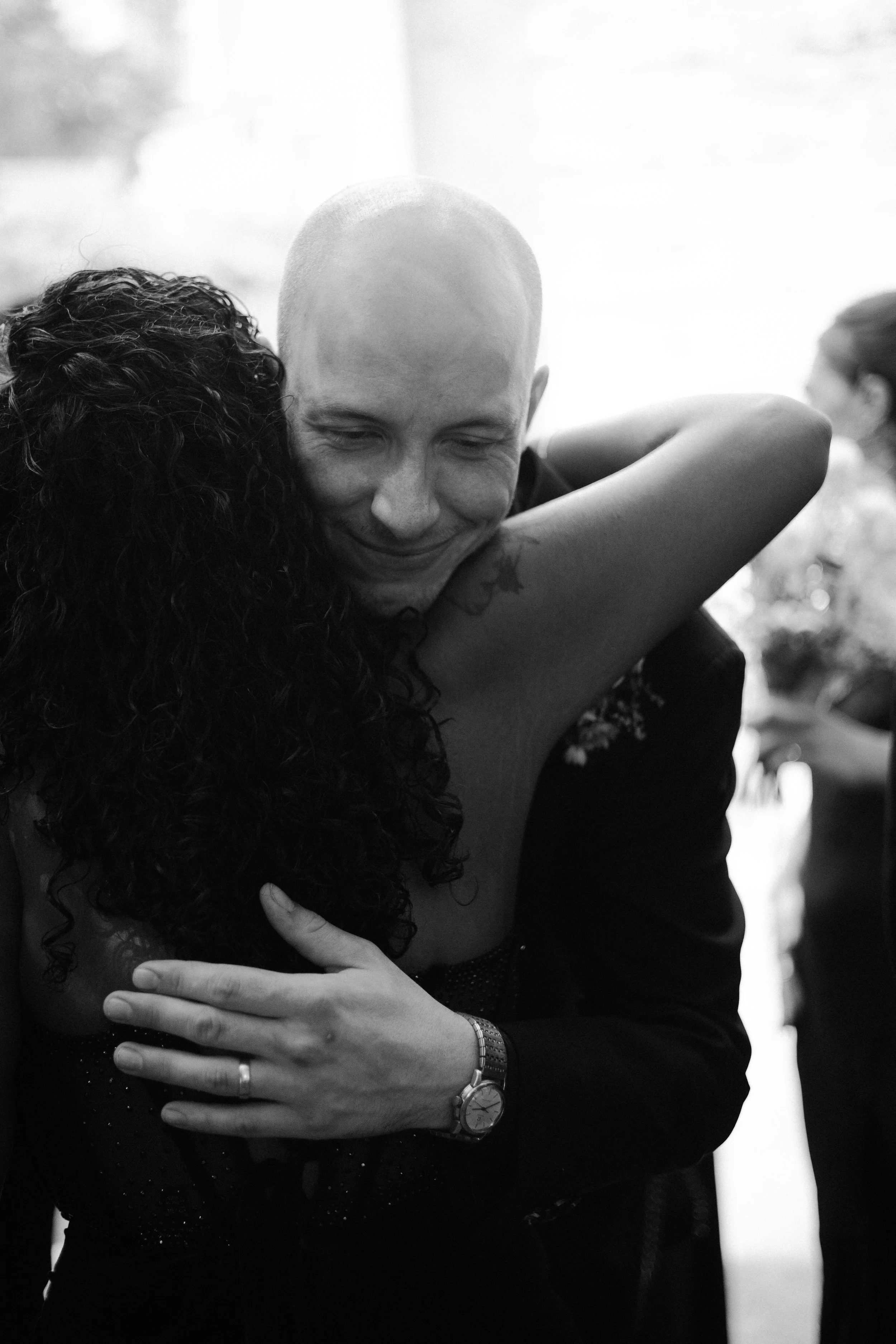 A person with a shaved head and tattoos hugging another person with curly hair, both embracing warmly in a close hug.