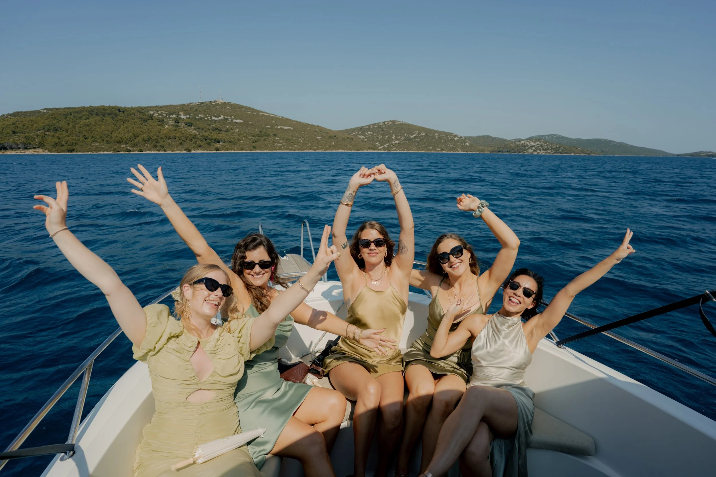 Five women wearing sunglasses and dresses enjoying a boat ride on the water with a hilly landscape in the background.