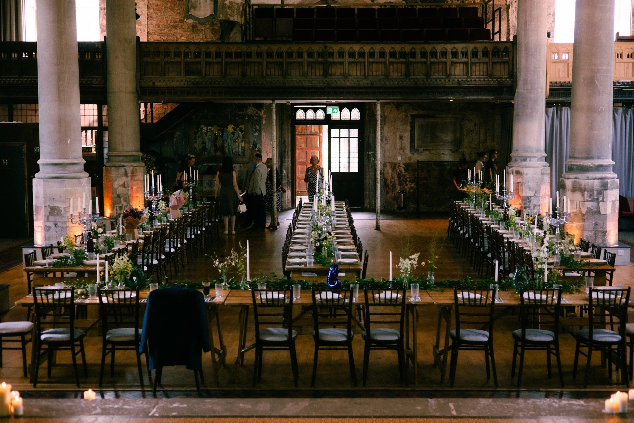 Indoor event space with long tables decorated with flowers and candelabras, tall white pillars, and a rustic brick wall background. Several people are standing and conversing near the entrance.