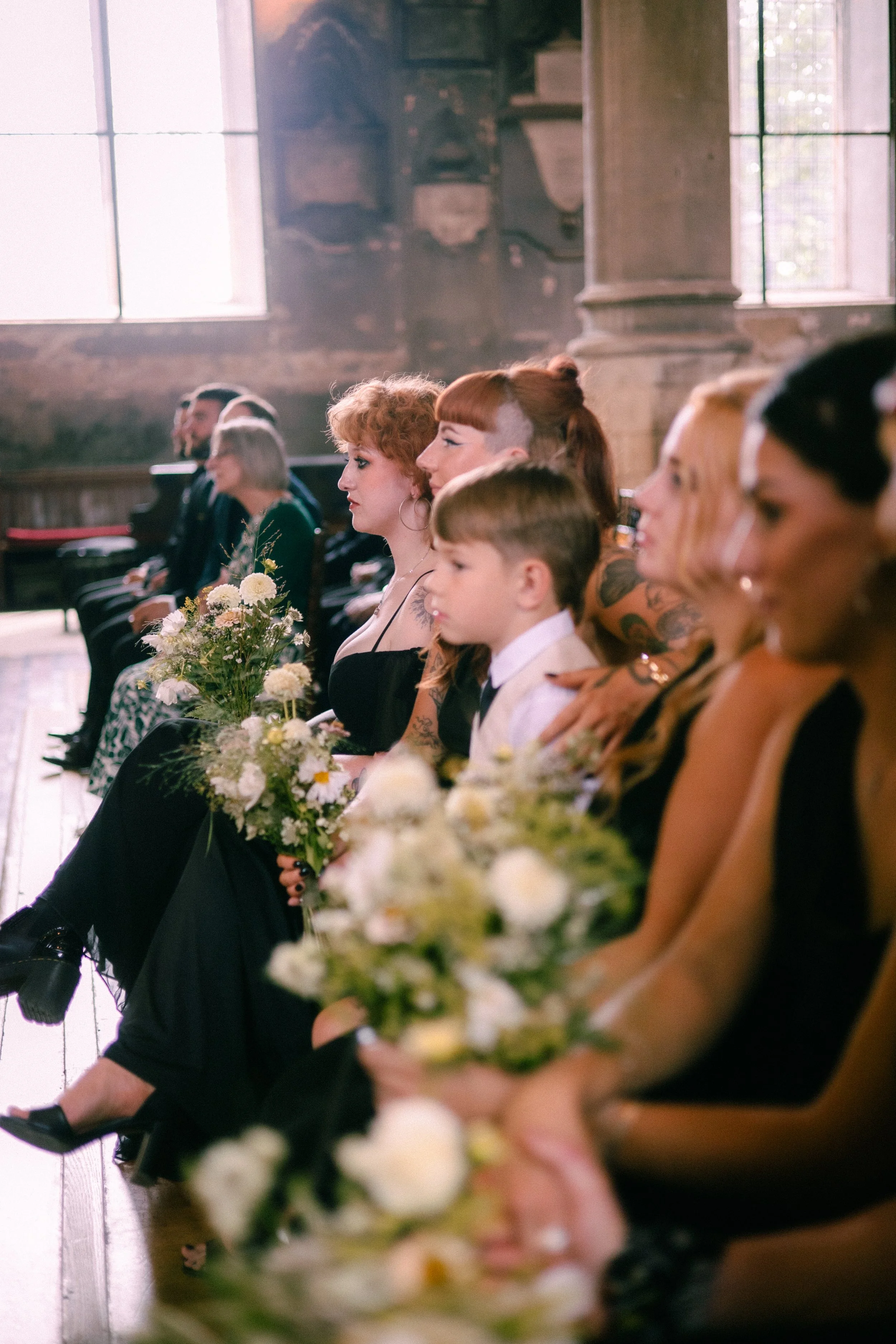 People sitting in a row at a formal event, holding bouquets of flowers, with sunlight coming through large windows in a historic stone building.
